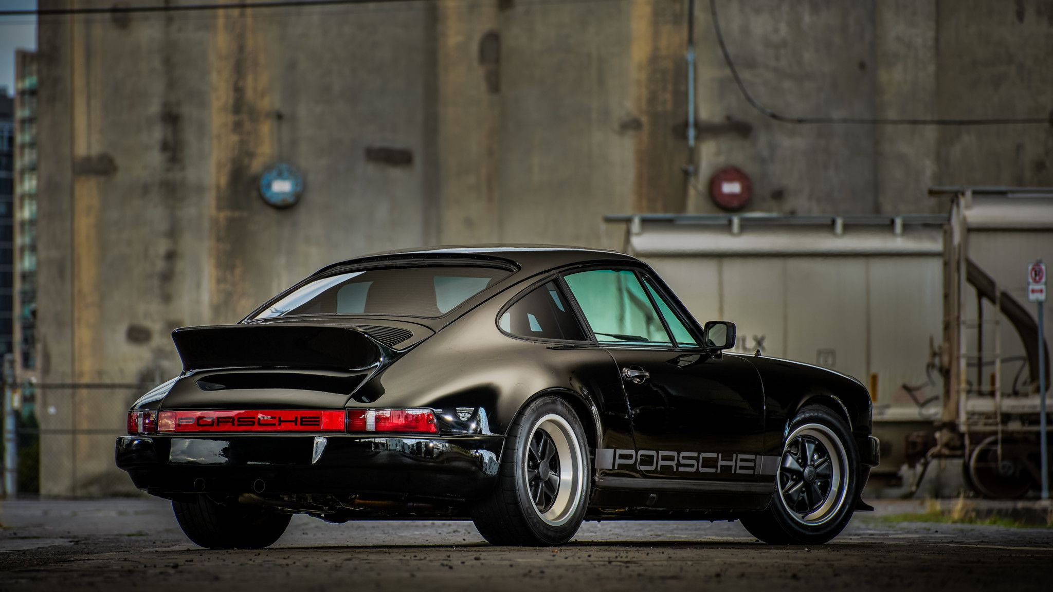 Porsche 911 桌布 壁紙 — 一輛時尚的黑色保時捷911 Turbo，帶有銀色條紋，停在風化的混凝土工業建築前。