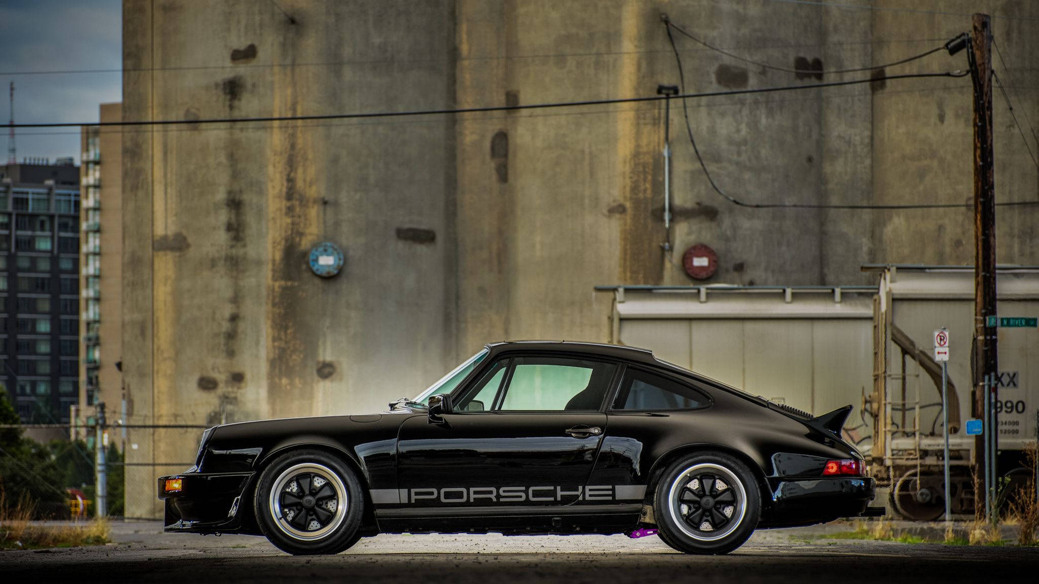Porsche 911 桌布 壁紙 — 一輛時尚的黑色保時捷911 Turbo停放在都市工業環境中風化的混凝土牆旁邊。