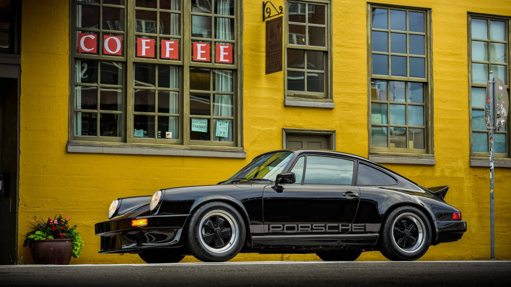 Porsche 911 桌布 壁紙 — 一輛時尚的黑色保時捷911停在充滿活力的黃色咖啡店前，帶有紅色字樣。