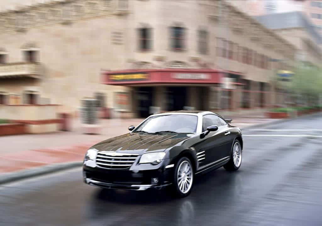 Chrysler Crossfire 桌布 壁紙 — 一輛時尚的黑色克萊斯勒Crossfire在濕滑的城市街道上疾馳，建築物呈現動態模糊，路面有倒影。