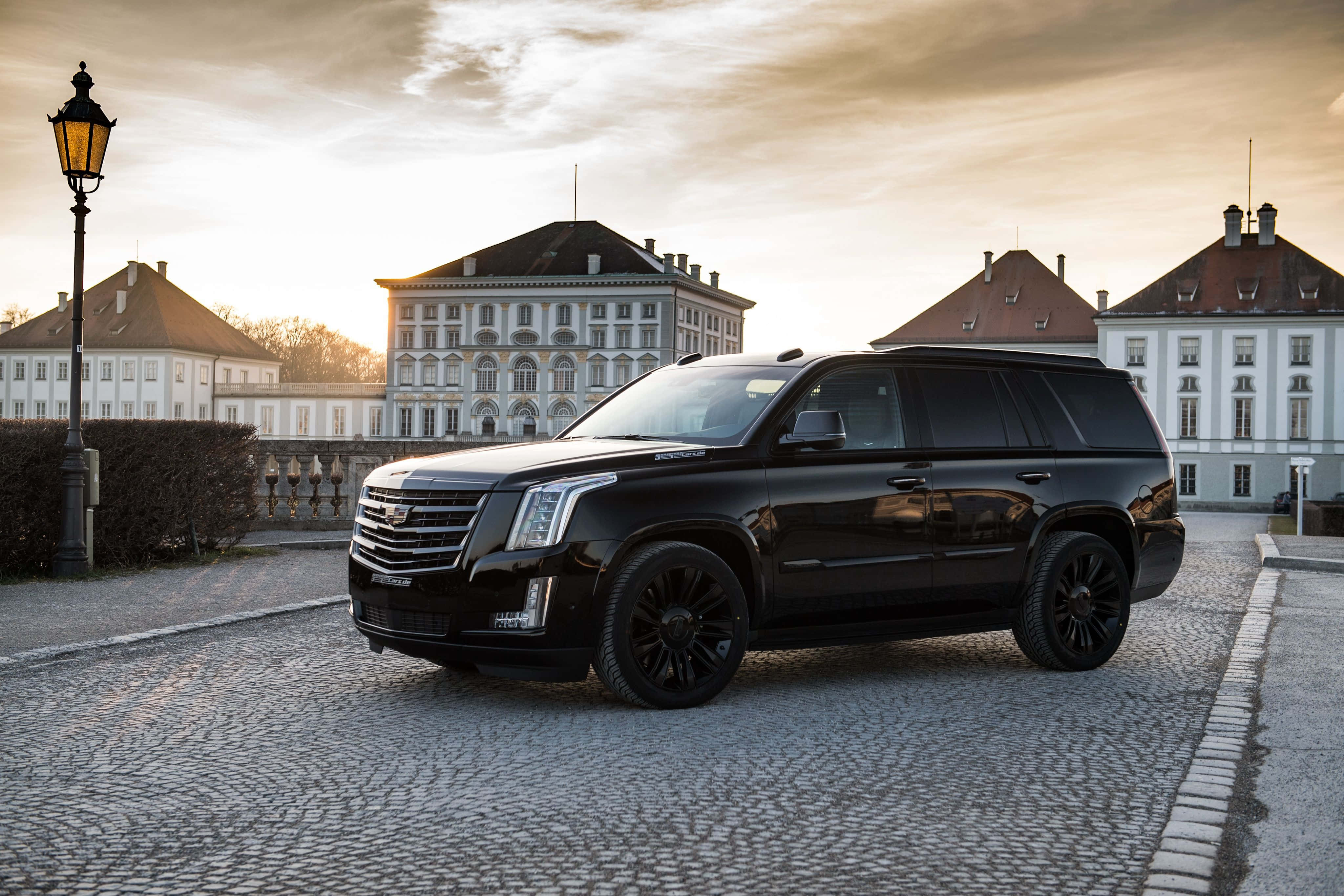 Cadillac Escalade 桌布 壁紙 — 一輛時尚的黑色凱迪拉克 Escalade 停泊在鵝卵石庭院中，前方是宏偉的歐式建築，正值黃金時刻。