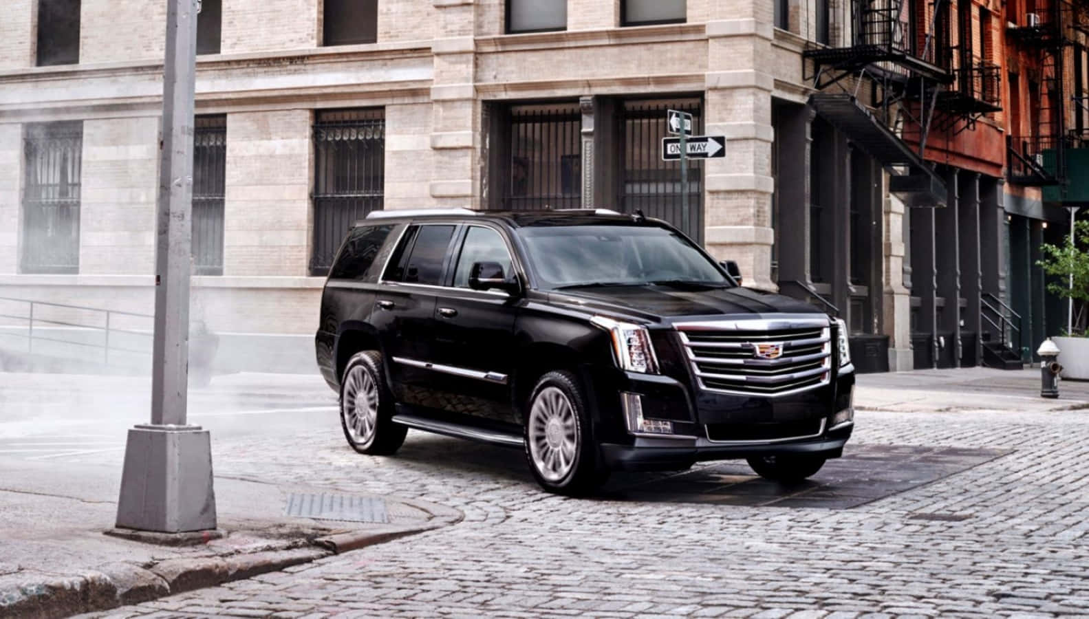 Cadillac Escalade 桌布 壁紙 — 一輛時尚的黑色凱迪拉克 Escalade 停放在濕漉漉的鵝卵石街道上，周圍有歷史建築和附近升起的蒸氣。