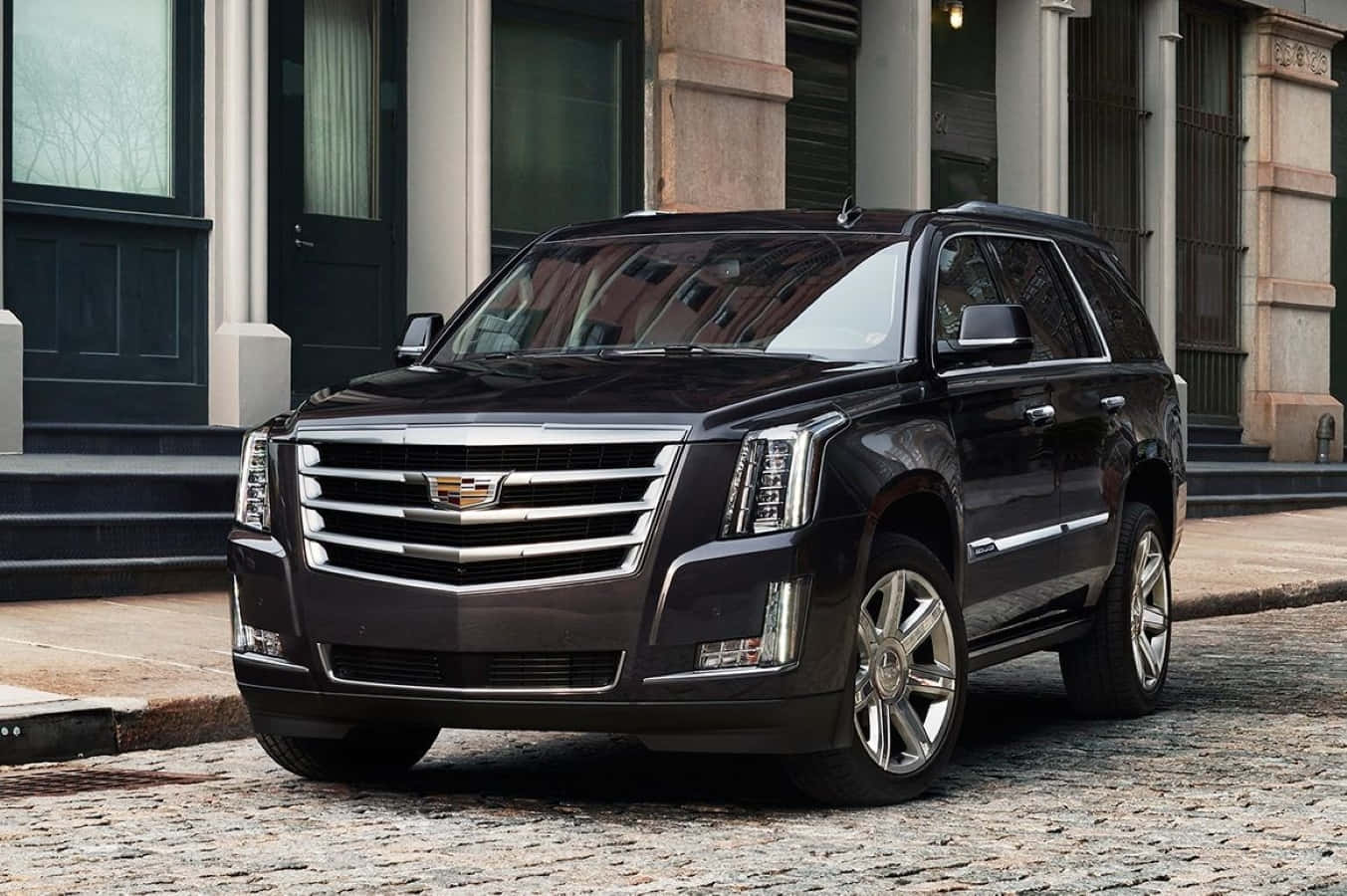 Cadillac Escalade 桌布 壁紙 — 一輛時尚的黑色凱迪拉克 Escalade SUV 停在鵝卵石街道上，前方是經典石造建築，帶有深色門。