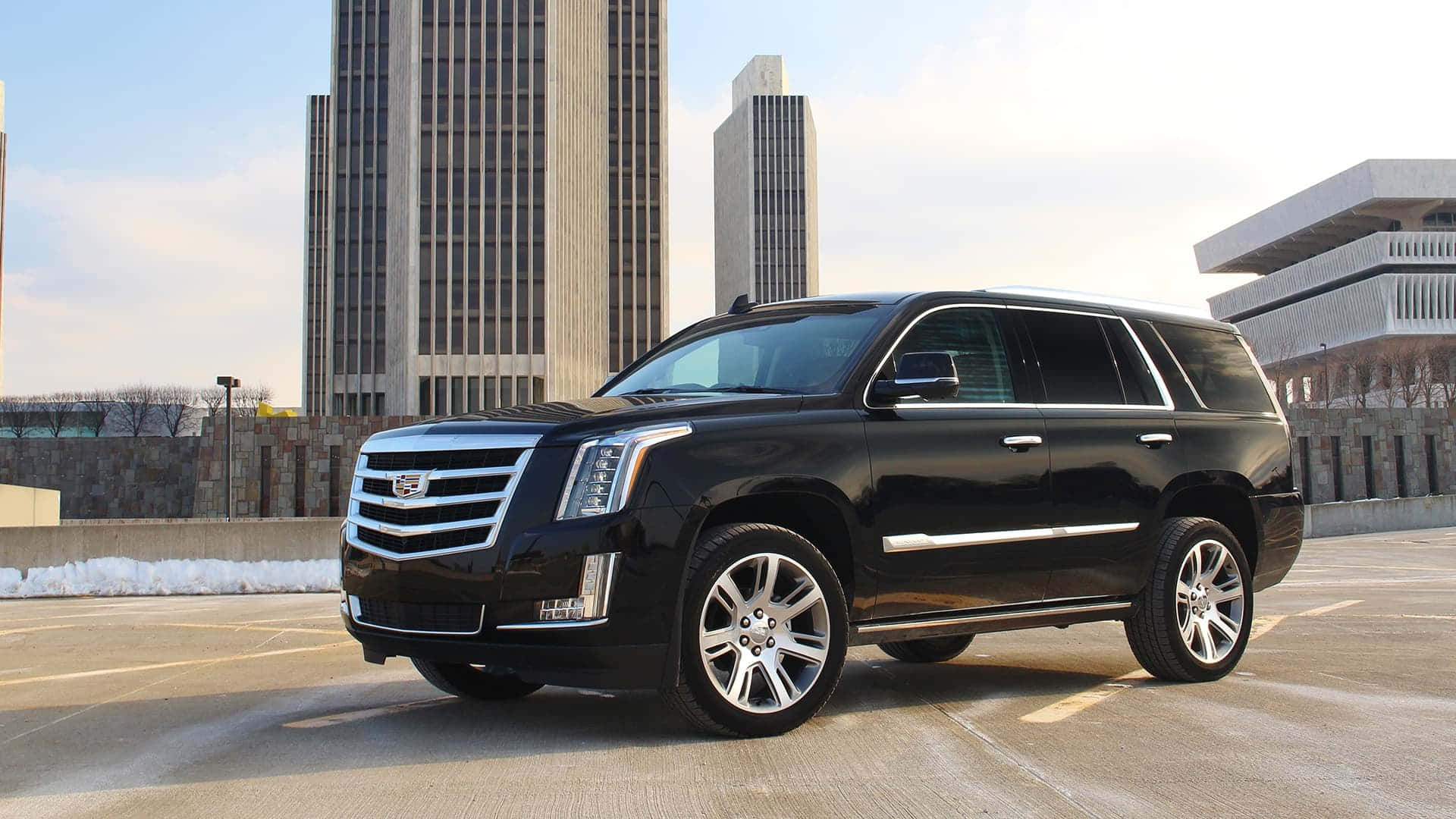 Cadillac Escalade 桌布 壁紙 — 一輛時尚的黑色凱迪拉克 Escalade SUV 停放在城市停車場，背景是現代摩天大樓和雪堆。