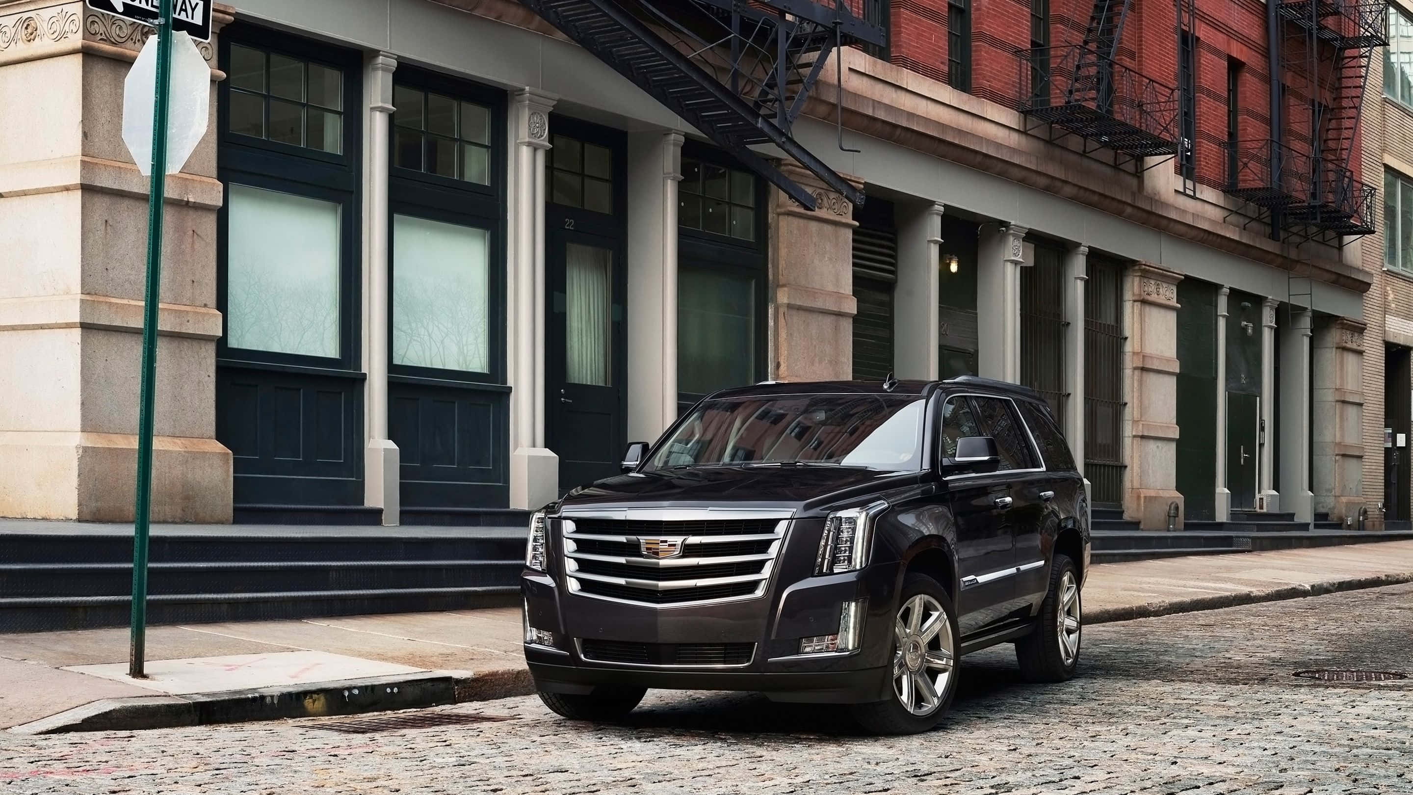 Cadillac Escalade 桌布 壁紙 — 一輛時尚的黑色凱迪拉克SUV停在鵝卵石街道上，前方是帶有防火梯的經典磚砌建築。