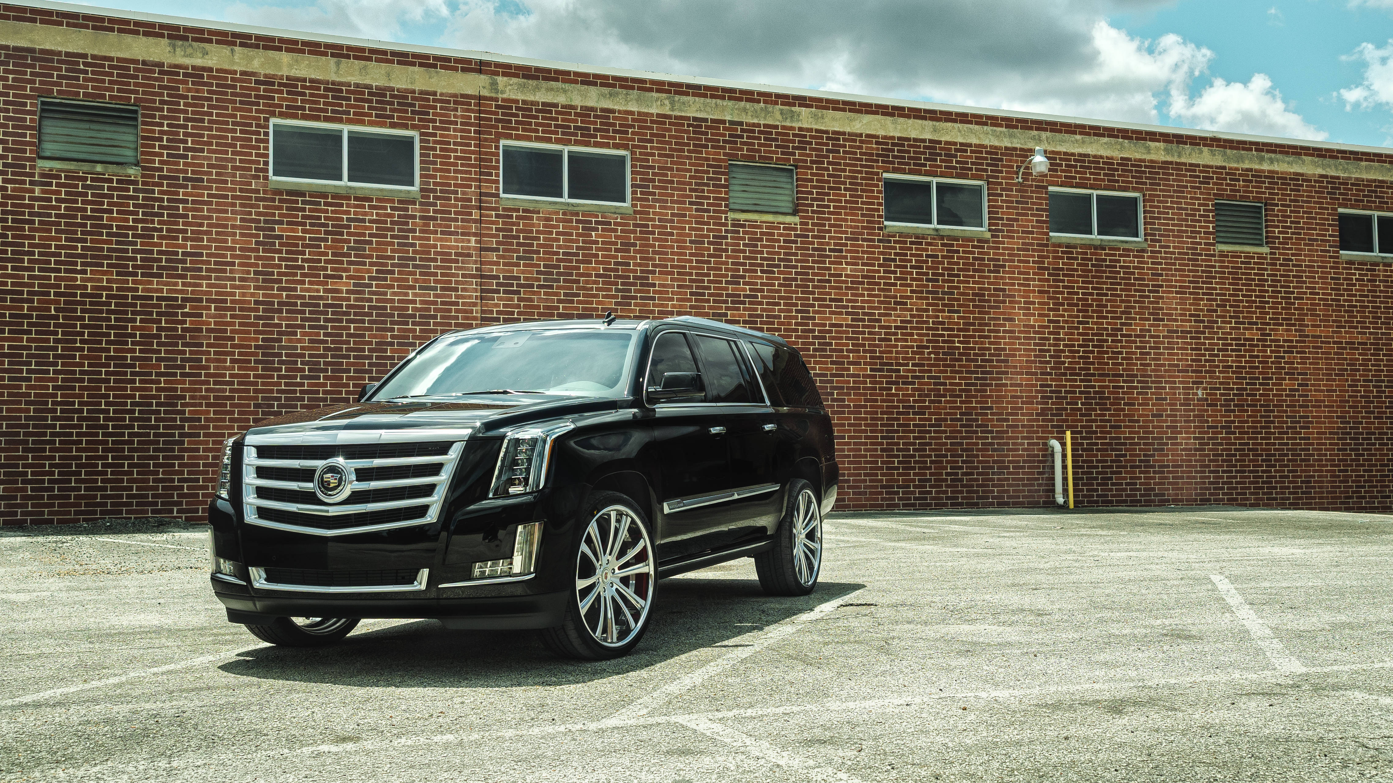 Cadillac 桌布 壁紙 — 一輛時尚的黑色凱迪拉克SUV停在陽光普照的混凝土停車場，背景是風化的磚砌建築，天空部分多雲。