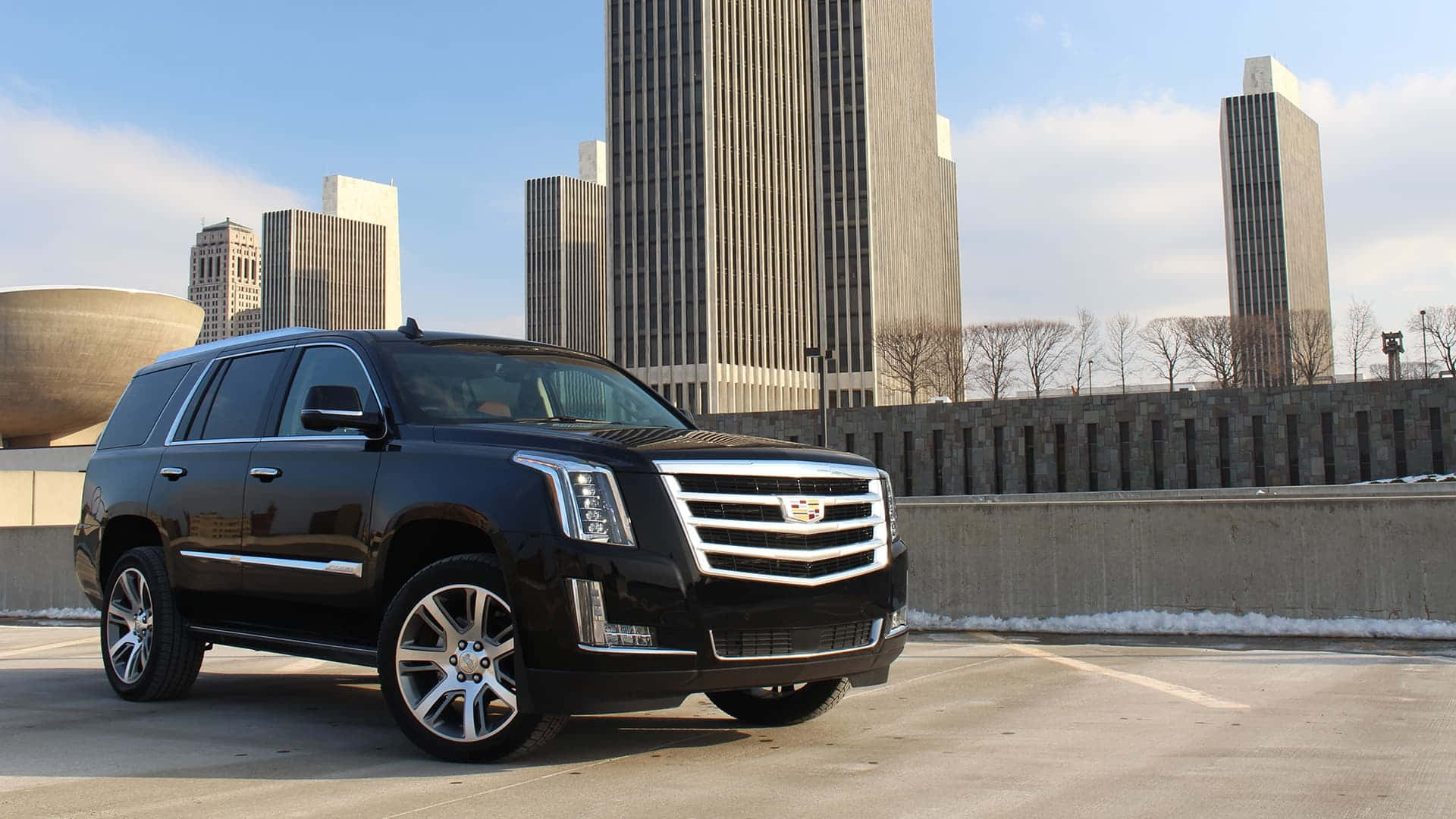 Cadillac Escalade 桌布 壁紙 — 一輛時尚的黑色凱迪拉克SUV停放在城市停車場，周圍有現代摩天大樓和光禿禿的樹木，天空湛藍。