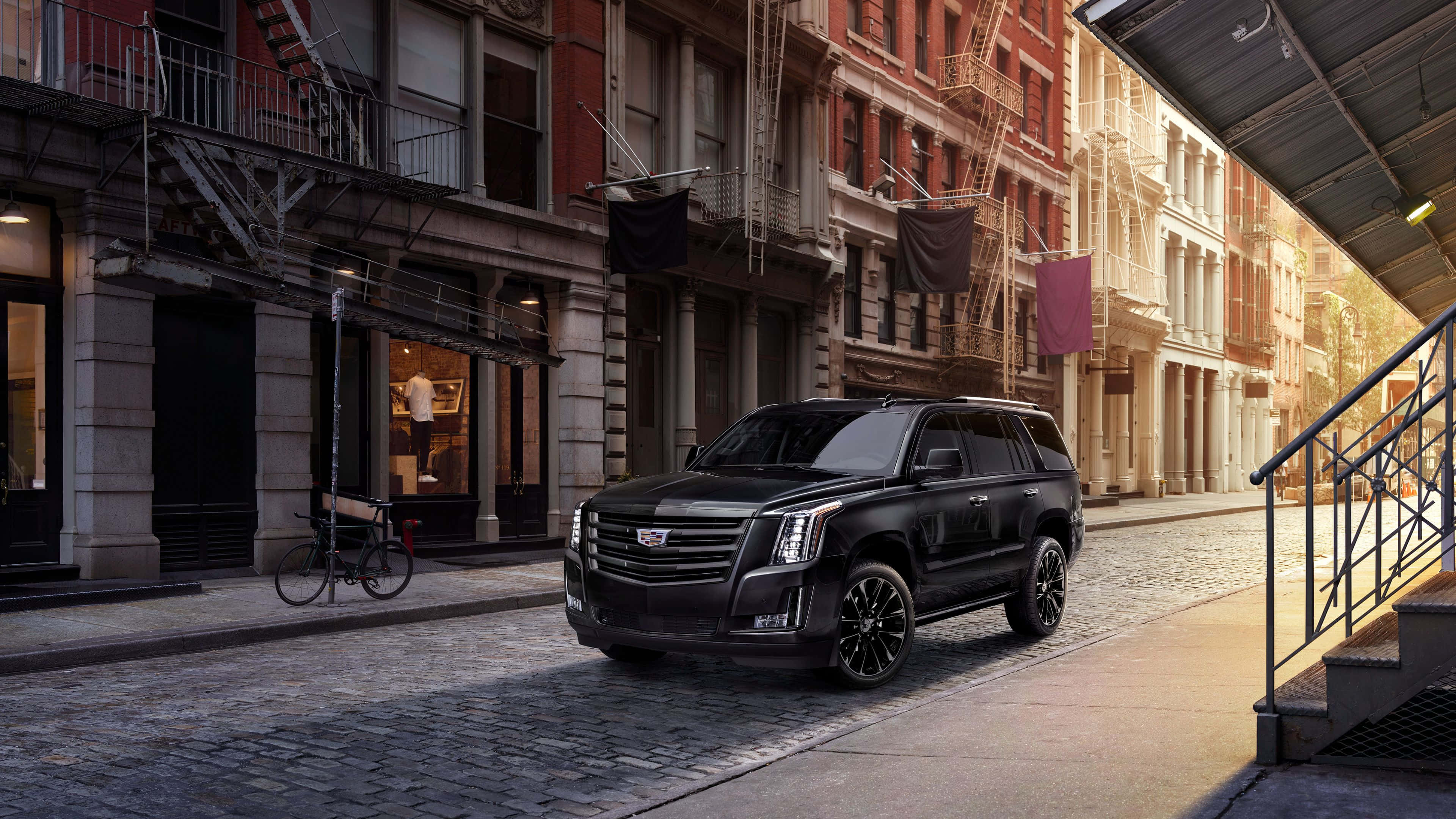 Cadillac Escalade 桌布 壁紙 — 一輛時尚的黑色凱迪拉克SUV停泊在陽光普照的鵝卵石街道上，兩旁是帶有防火逃生梯的經典磚砌建築。