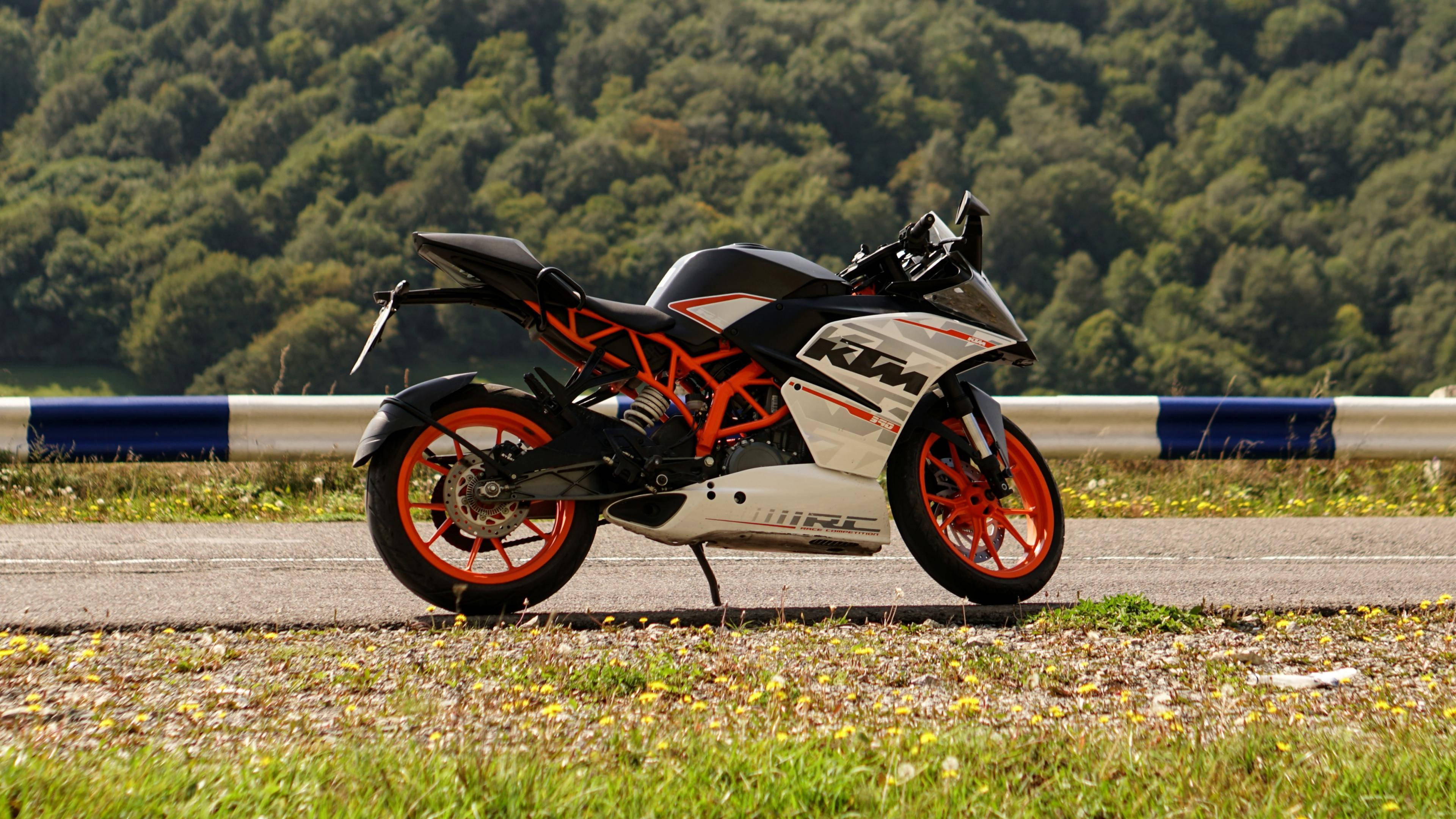 KTM 摩托車 桌布 壁紙 — 一輛時尚的黑色和橙色KTM機車停放在路邊，背景是樹木繁茂的山丘。