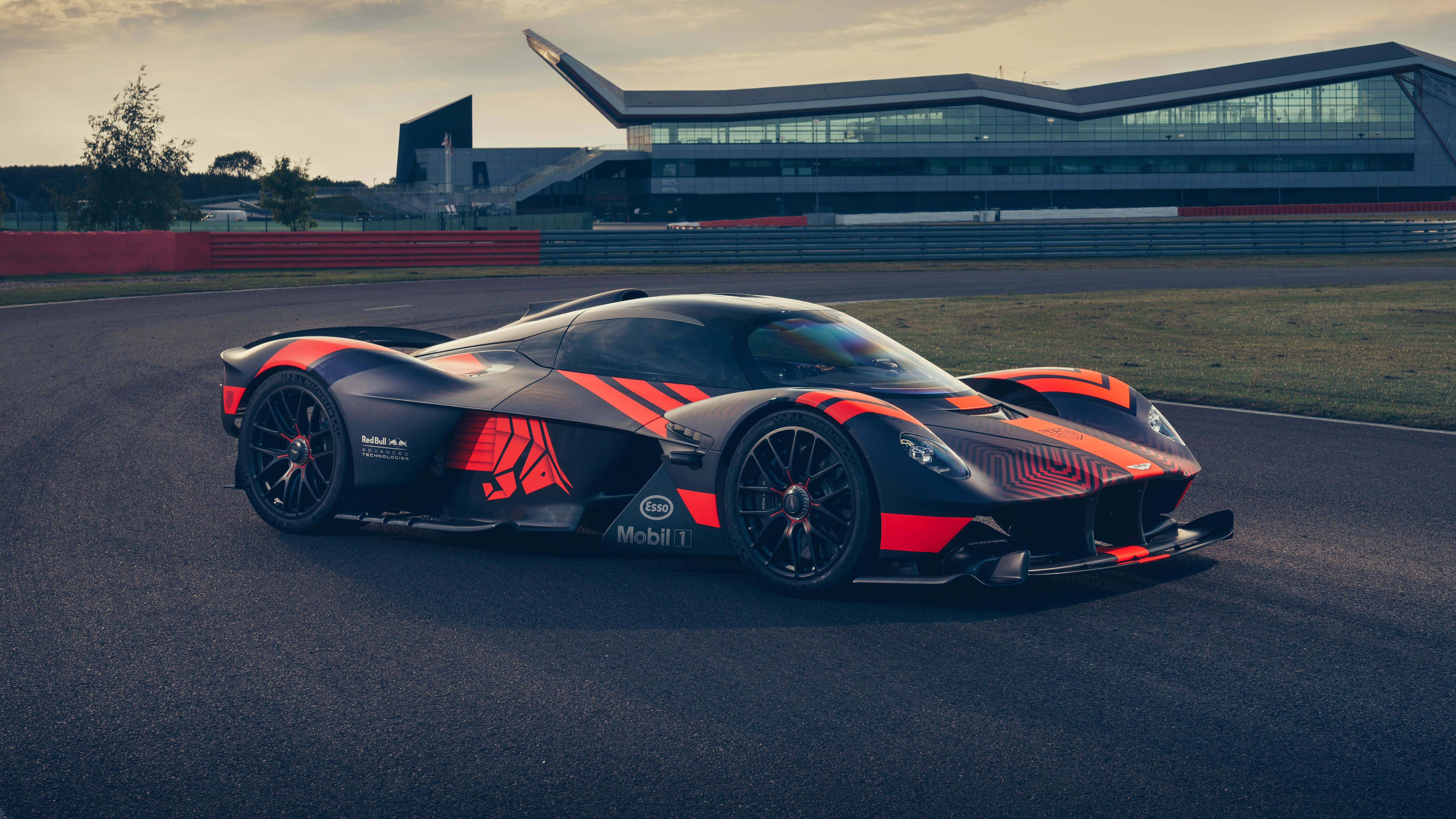 Aston Martin Valkyrie 桌布 壁紙 — 一輛時尚的黑色和橙色超跑帶有醒目的紅色點綴，停在賽道上，旁邊是一座現代玻璃建築，背景是柔和的暮色天空。