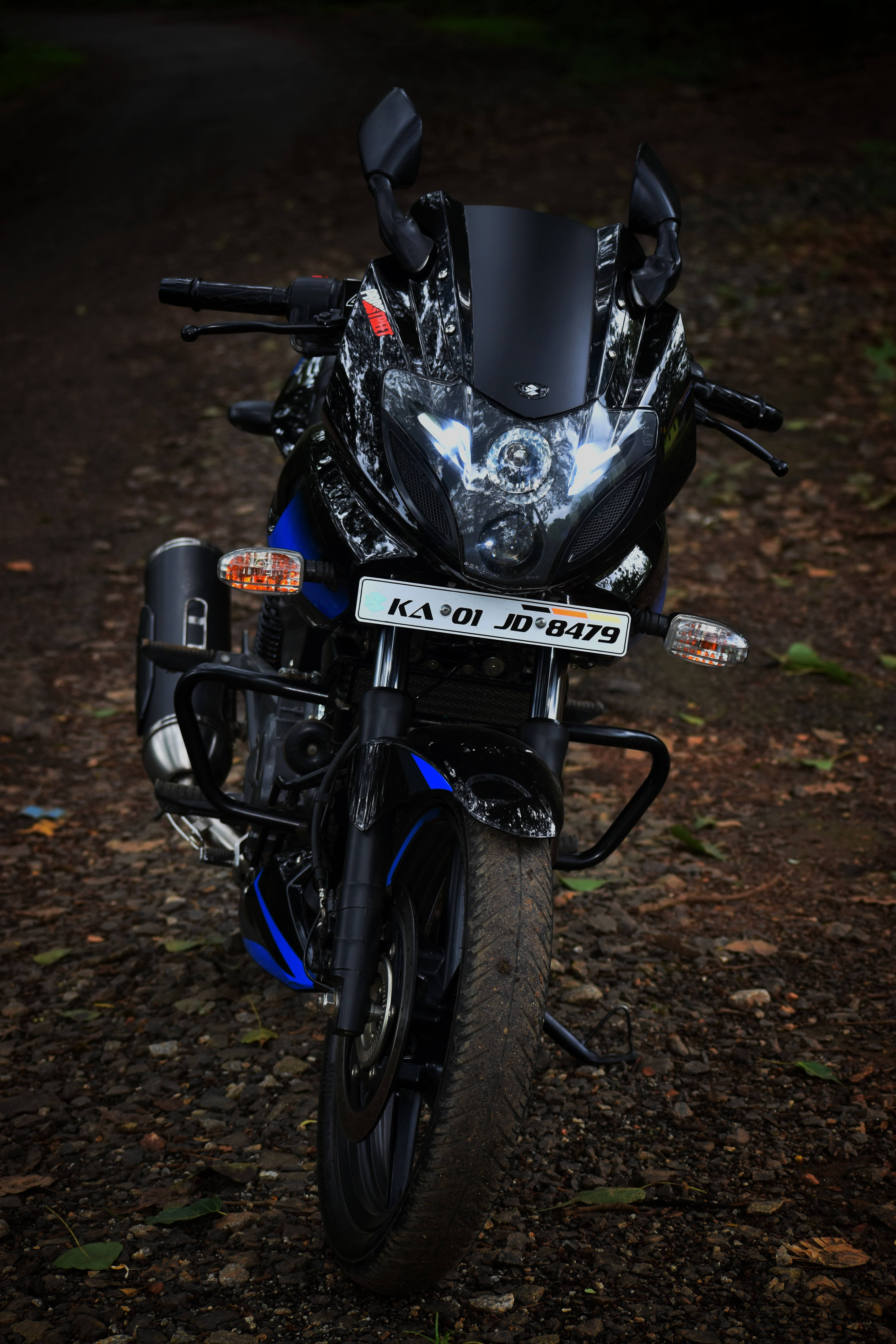 Pulsar 220 HD 桌布 壁紙 — 一輛時尚的黑色和藍色運動機車，配備發光的頭燈，在昏暗的森林光線下停在碎石路上。