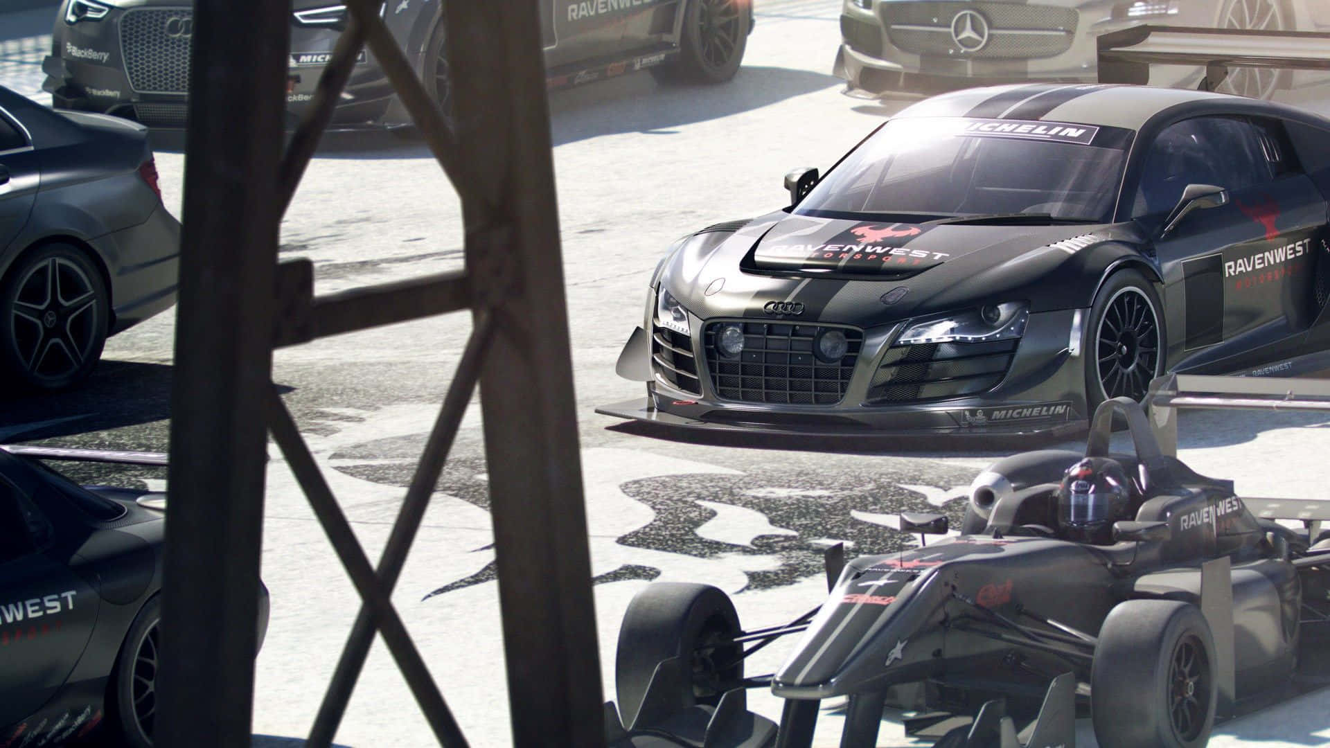 1080p Grid Autosport 桌布 壁紙 — 一輛時尚的黑色奧迪R8賽車和一輛一級方程式賽車停放在陽光明媚的維修區車道上，其他高性能車輛也清晰可見。