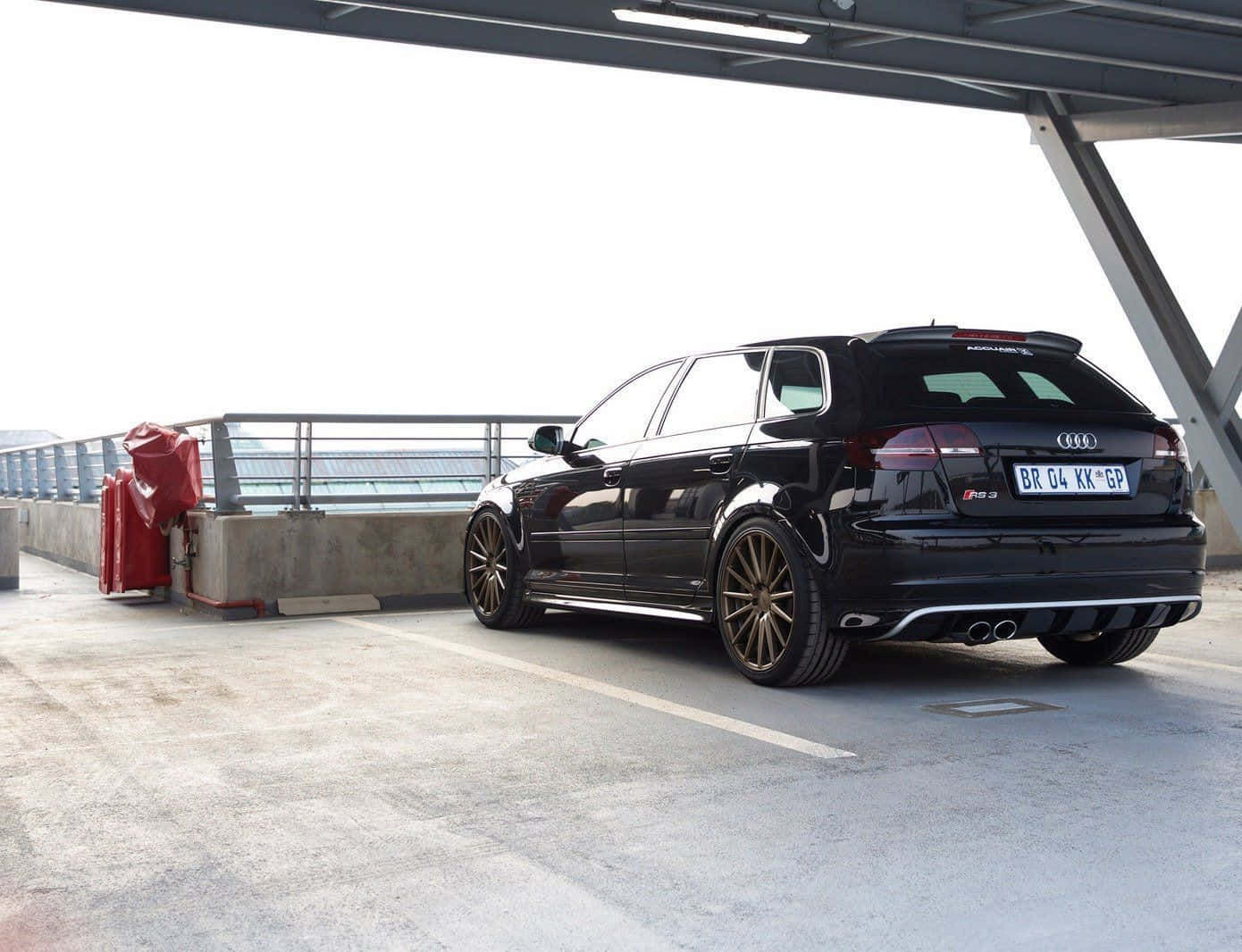 Audi Rs3 桌布 壁紙 — 一輛時尚的黑色奧迪RS3 Sportback停泊在現代鋼橋下，配備金色輪圈和雙出排氣管。