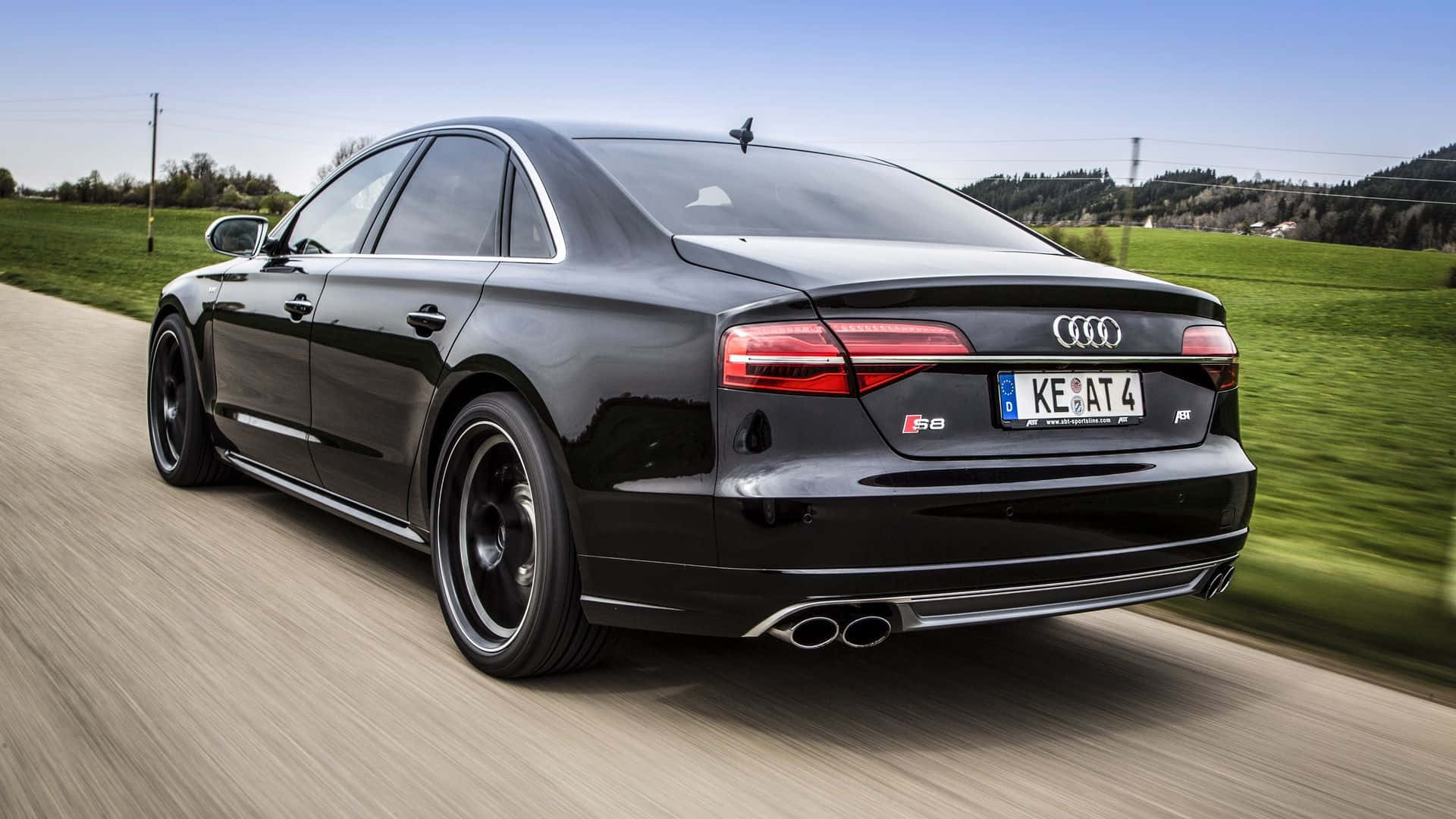 Audi S8 桌布 壁紙 — 一輛時尚的黑色奧迪S8轎車在鄉村道路上奔馳，背景是模糊的綠色田野和山丘，頭頂是清澈的藍天。