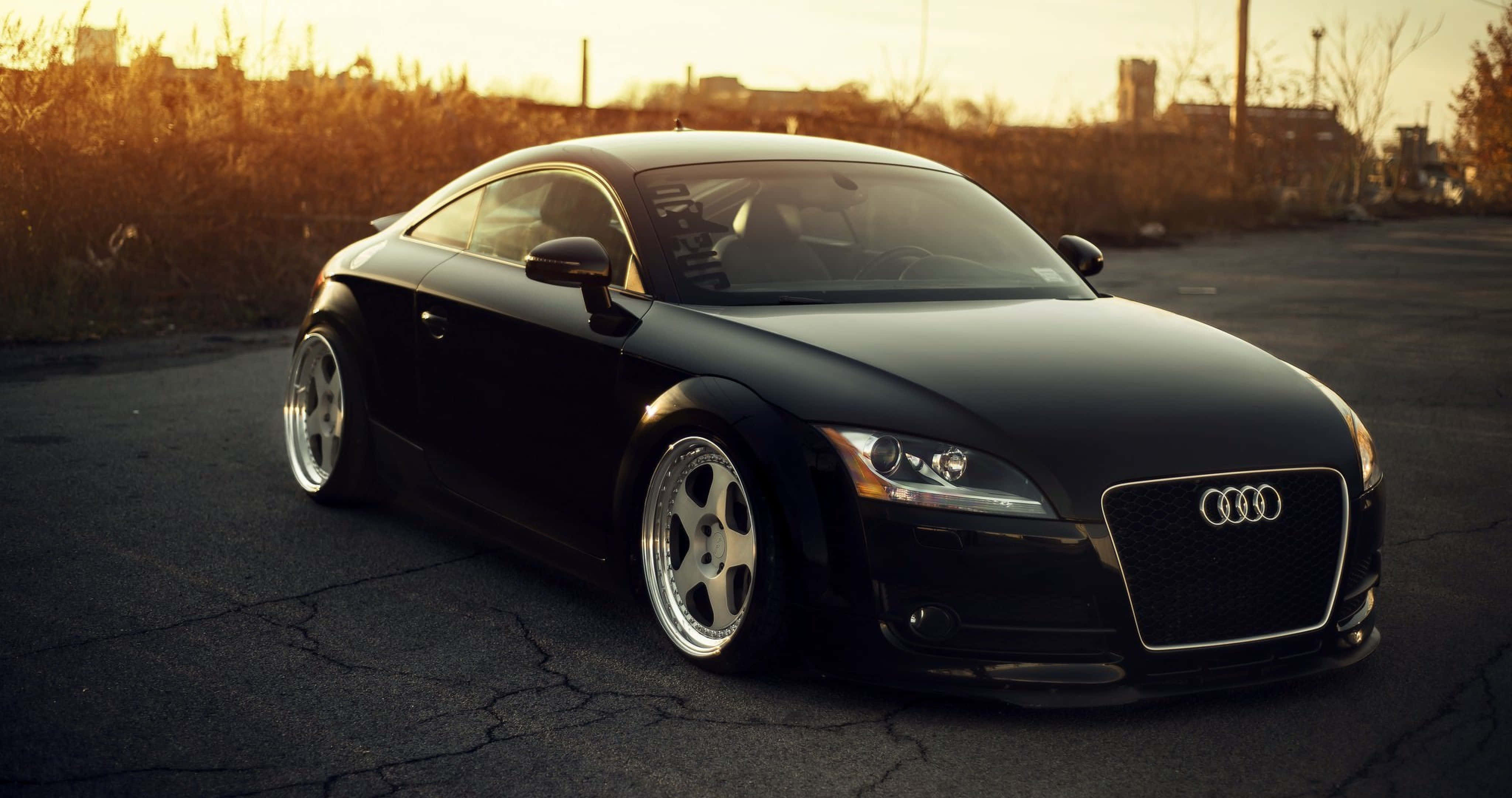 Audi Tt 桌布 壁紙 — 一輛時尚的黑色奧迪TT雙門跑車停泊在龜裂的柏油路上，沐浴在溫暖的金色夕陽光中。