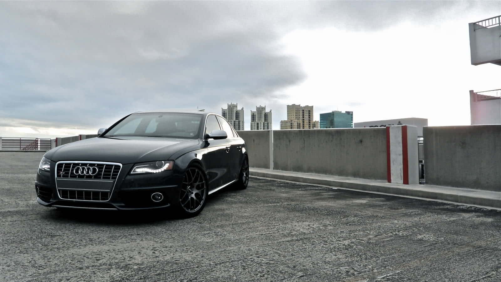 Audi A4 桌布 壁紙 — 一輛時尚的黑色奧迪房車停泊在一個佈滿碎石的天台停車場，天空陰沉，遠處有城市建築。