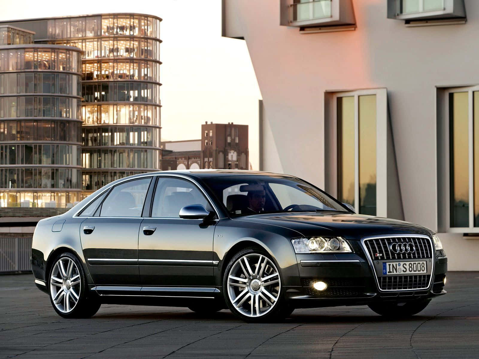 Audi S8 桌布 壁紙 — 一輛時尚的黑色奧迪轎車停在石頭廣場上，夕陽西下，背後現代玻璃建築閃閃發光。