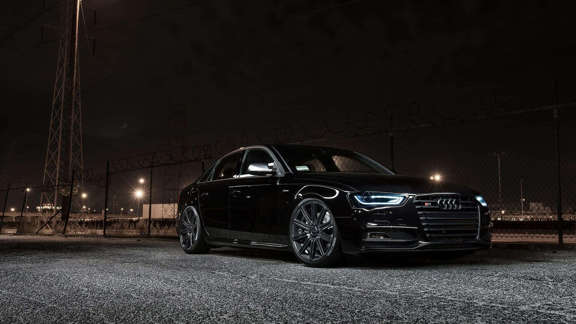 Audi S4 桌布 壁紙 — 一輛時尚的黑色奧迪轎車在夜燈下閃閃發光，停在帶有鐵絲網的鐵鏈圍欄旁。