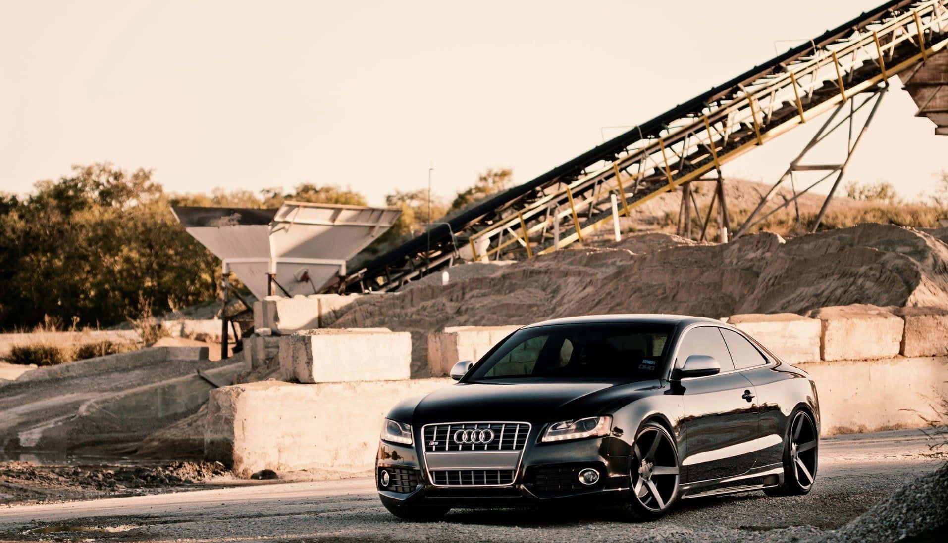 Audi S6 桌布 壁紙 — 一輛時尚的黑色奧迪雙門轎跑車停在碎石上，靠近工業混凝土塊和輸送帶，沐浴在溫暖的金色光線中。