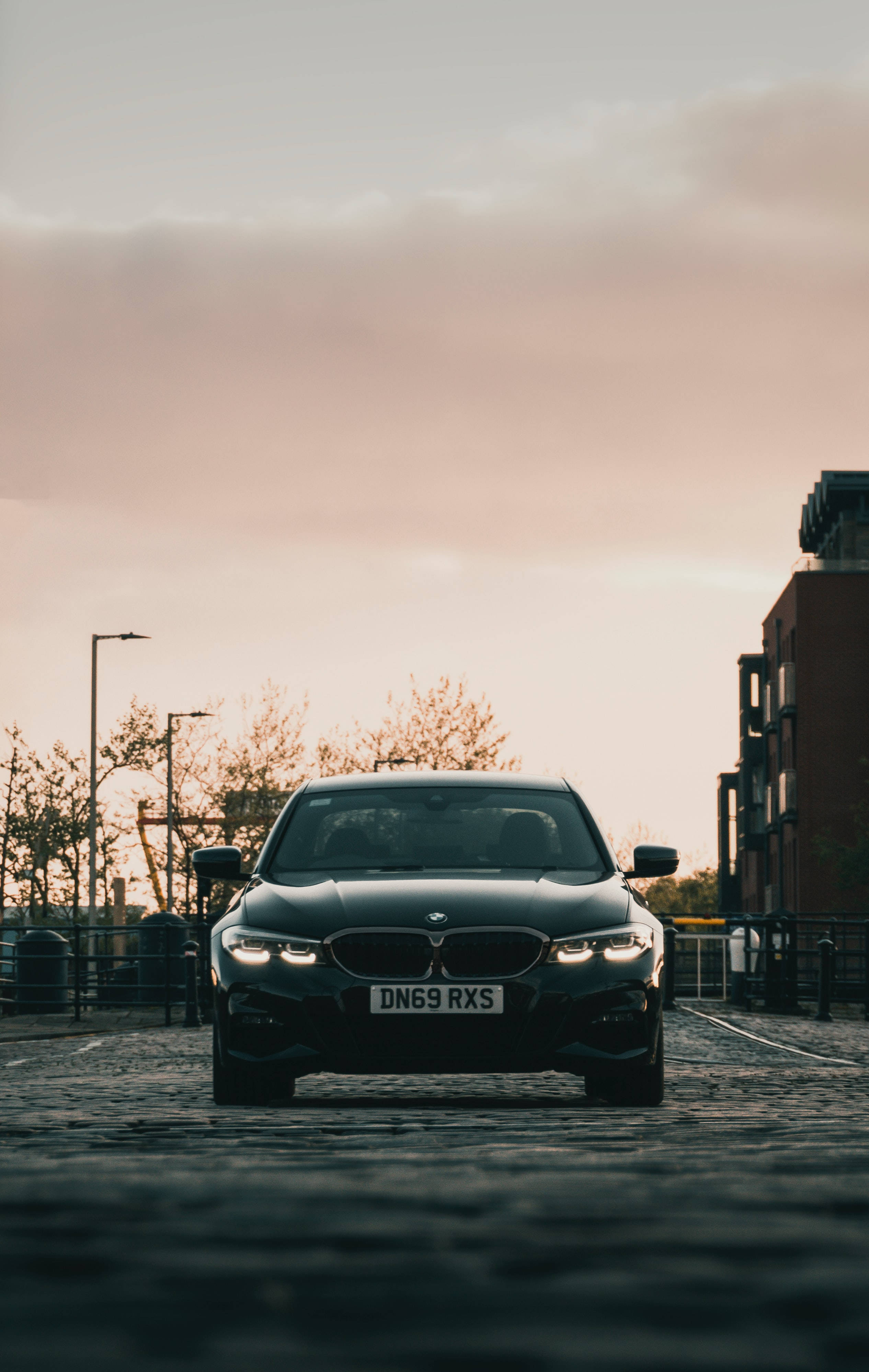 Bmw M Series 桌布 壁紙 — 一輛時尚的黑色寶馬轎車在日落時分朝前停在鵝卵石街道上，周圍有溫暖的環境照明。