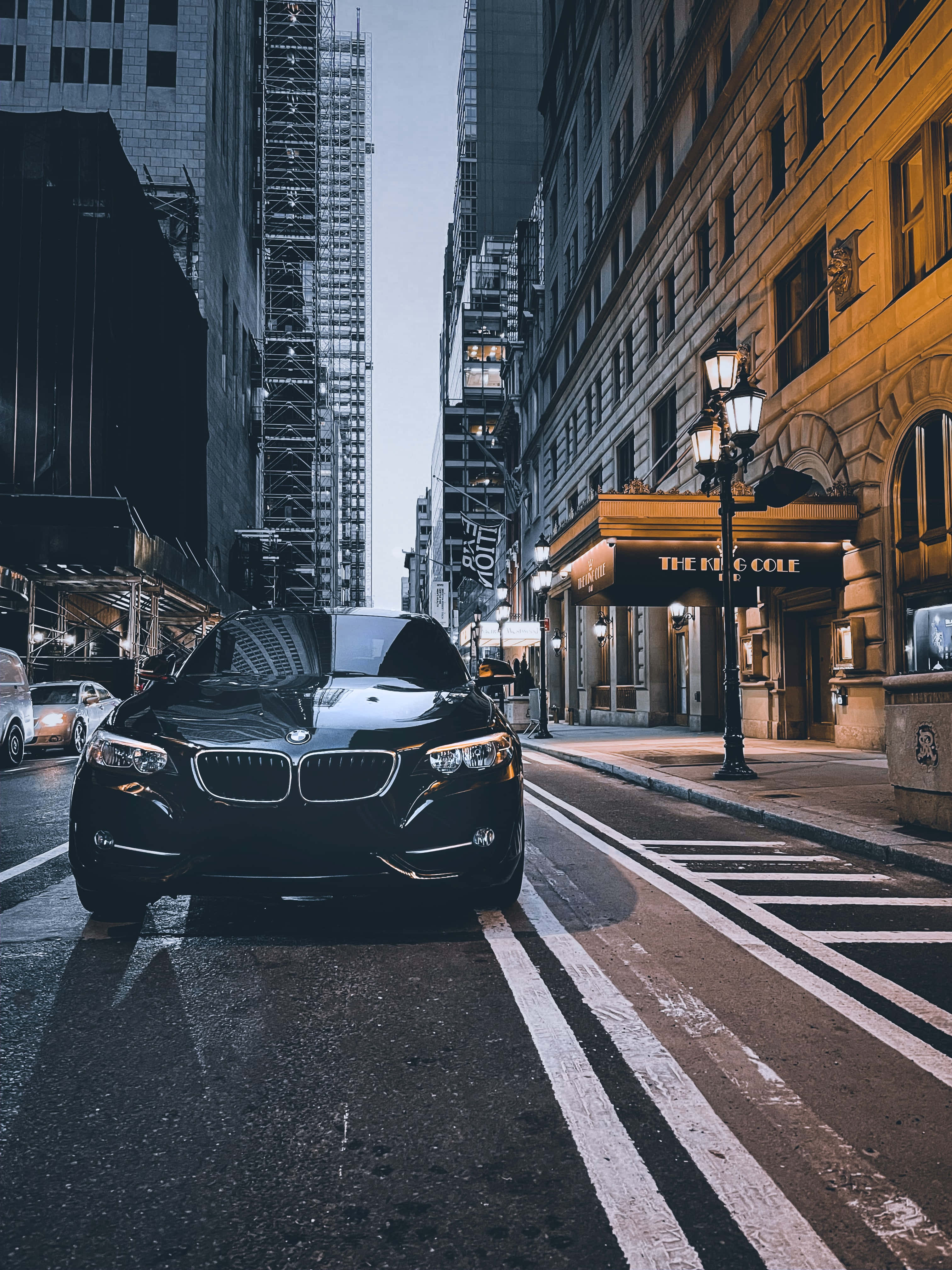 BMW 動圖 桌布 壁紙 — 一輛時尚的黑色寶馬轎車在濕漉漉的城市街道上向前行駛，兩旁是高聳的建築物和燈火通明的飯店入口。