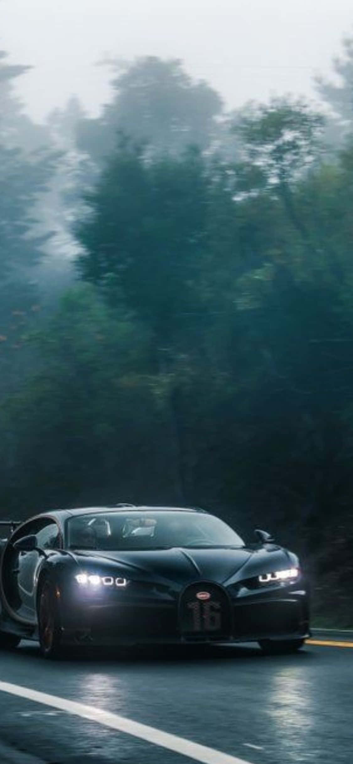 iPhone Xs Bugatti 桌布 壁紙 — 一輛時尚的黑色布加迪Chiron在潮濕的森林道路上奔馳，周圍是霧氣籠罩的綠樹，車頭燈閃閃發光。