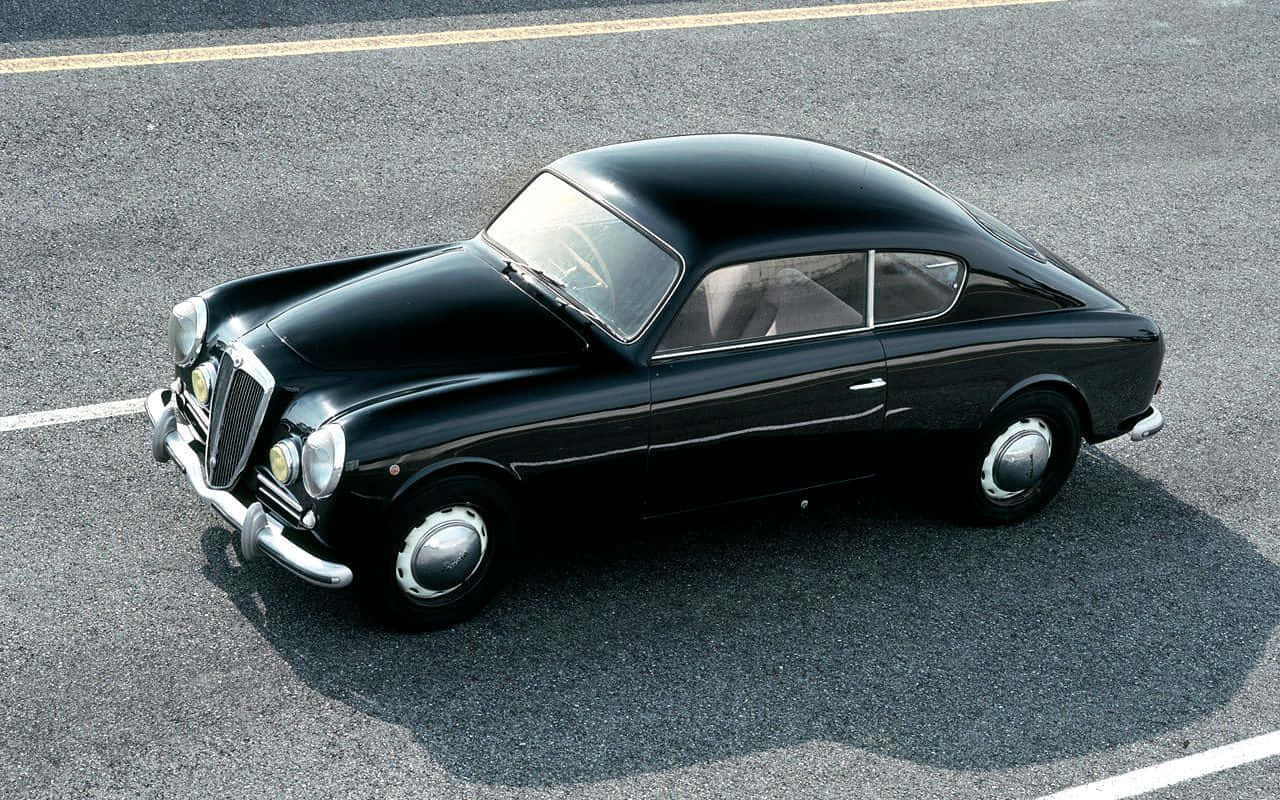 Lancia 桌布 壁紙 — 一輛時尚的黑色復古跑車，帶有鍍鉻裝飾，停在明亮的陽光下的鋪砌停車場上。