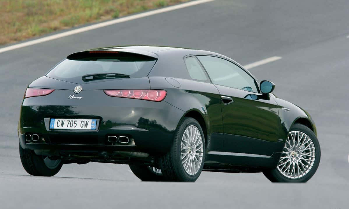 Alfa Romeo Brera 桌布 壁紙 — 一輛時尚的黑色愛快羅密歐 Brera 跑車沿著蜿蜒的瀝青道路行駛，背景是模糊的綠色植物。