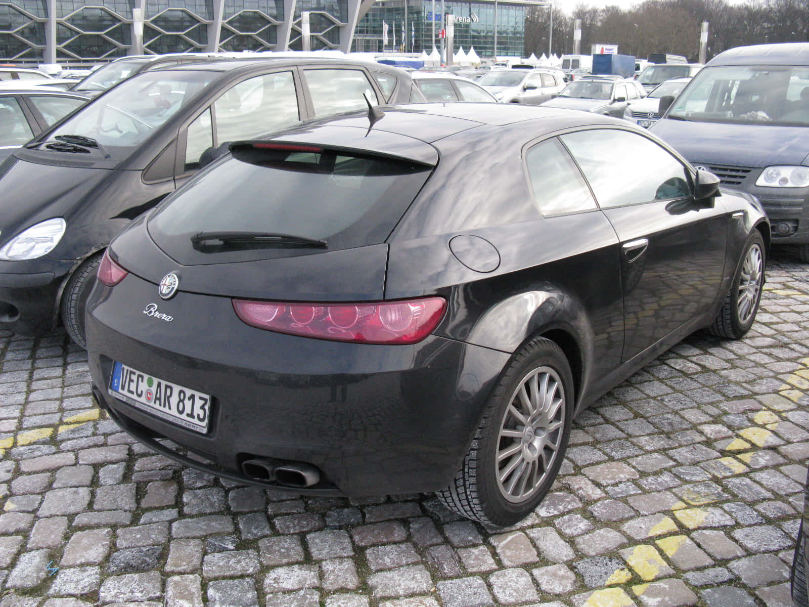 Alfa Romeo Brera 桌布 壁紙 — 一輛時尚的黑色愛快羅密歐 Brera 雙門跑車停放在鵝卵石停車場，背景是現代建築。