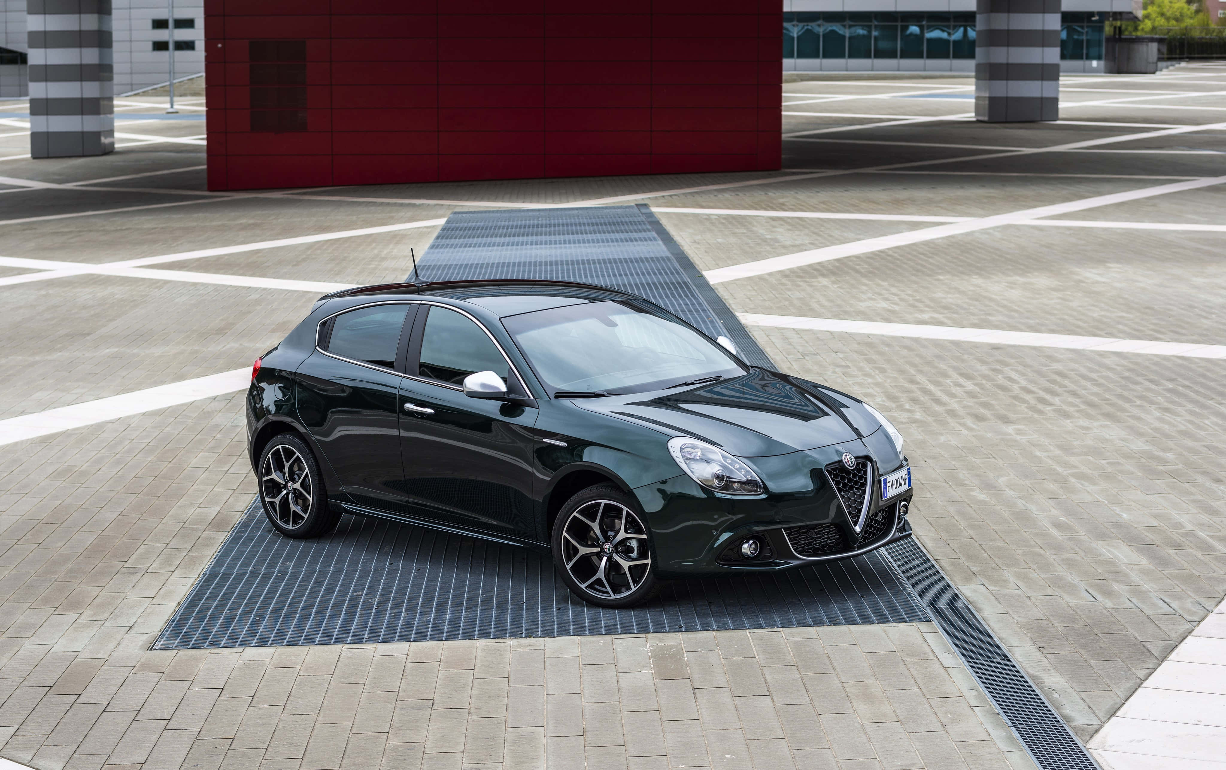Alfa Romeo Giulietta 桌布 壁紙 — 一輛時尚的黑色愛快羅密歐 Giulietta 停放在現代鋪砌廣場上，背景是鮮明的紅色建築。