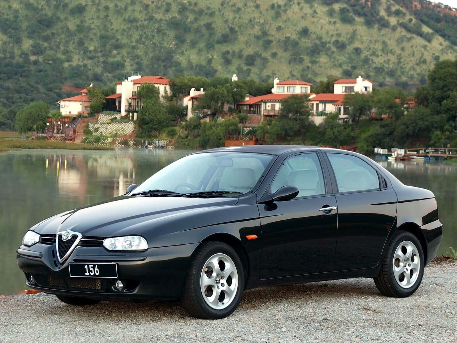 Alfa Romeo 156 桌布 壁紙 — 一輛時尚的黑色愛快羅密歐156轎車停在平靜湖泊旁的碎石地上，背景是山坡上的房屋。