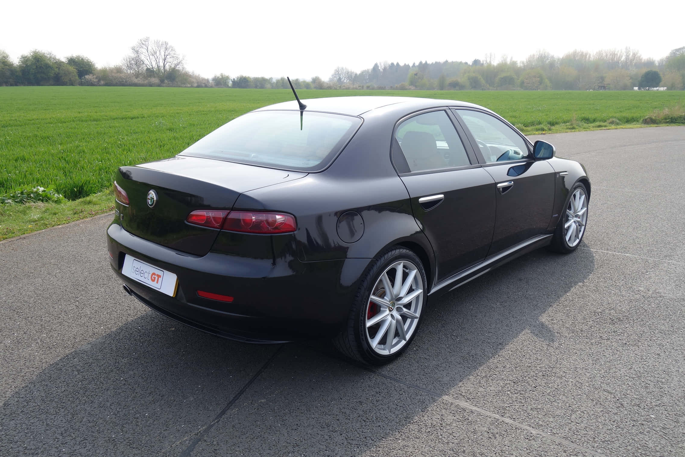 Alfa Romeo 159 桌布 壁紙 — 一輛時尚的黑色愛快羅密歐GT轎車停在鋪砌的道路上，旁邊是充滿活力的綠色田野，天空明亮。