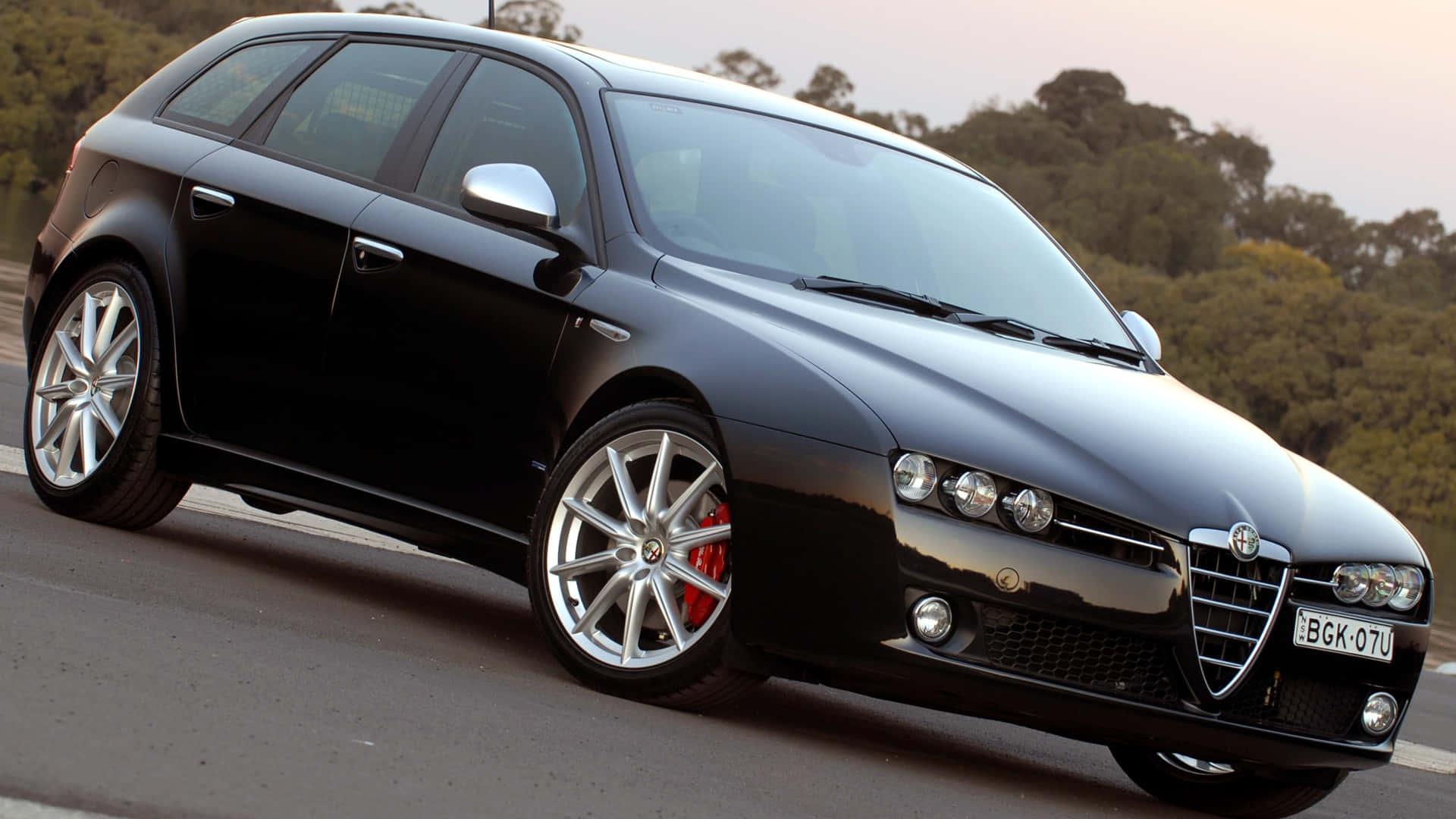 Alfa Romeo 159 桌布 壁紙 — 一輛時尚的黑色愛快羅密歐旅行車，配備銀色輪圈和紅色制動卡鉗，在黃昏時停泊在道路上，背景有樹木。