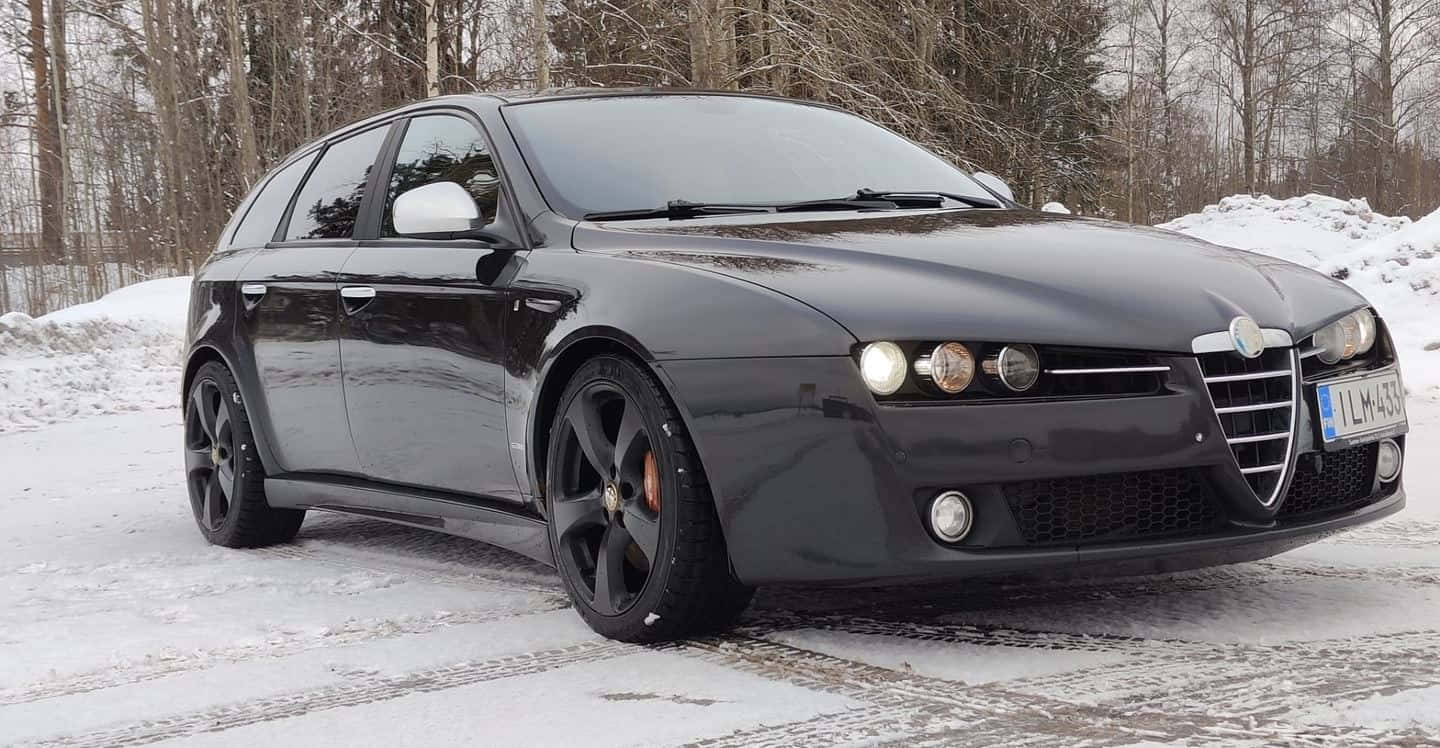 Alfa Romeo 159 桌布 壁紙 — 一輛時尚的黑色愛快羅密歐旅行車停放在積雪的森林道路上，背景是光禿的樹木。
