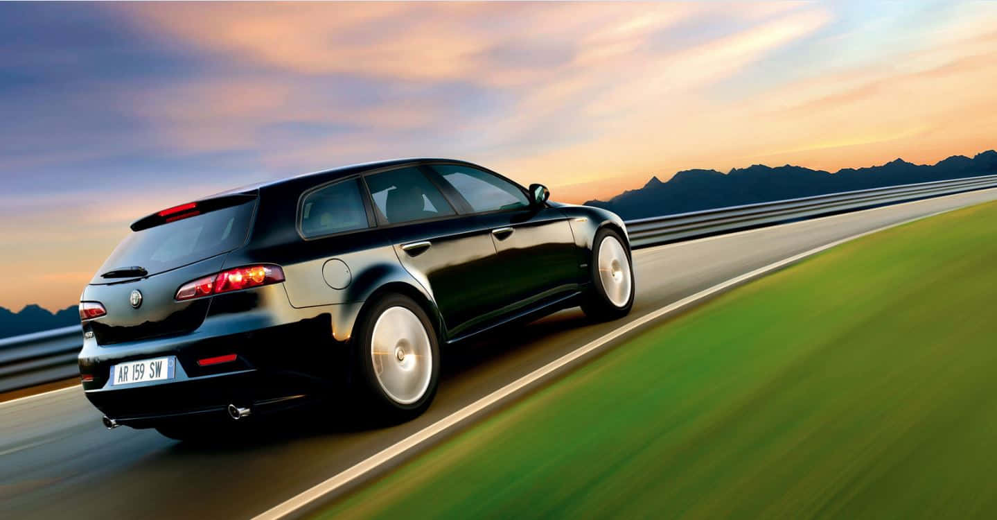 Alfa Romeo 159 桌布 壁紙 — 一輛時尚的黑色愛快羅密歐旅行車在日落時分沿著蜿蜒的高速公路疾馳，背景是模糊的綠色田野和遠山。
