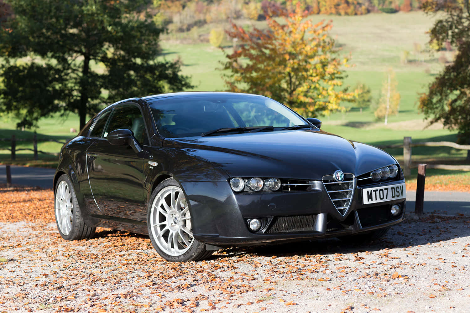 Alfa Romeo Brera 桌布 壁紙 — 一輛時尚的黑色愛快羅密歐跑車停在碎石路上，周圍是秋葉，背景是起伏的綠色山丘和色彩繽紛的樹木。