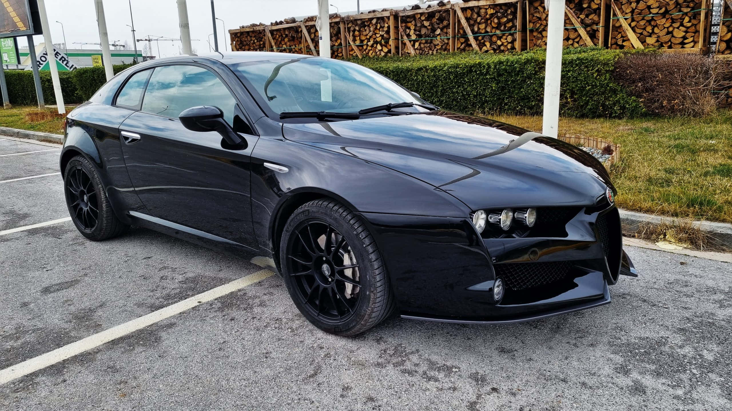 Alfa Romeo 159 桌布 壁紙 — 一輛時尚的黑色愛快羅密歐跑車停放在停車場，背景有堆疊木材和綠色樹籬。