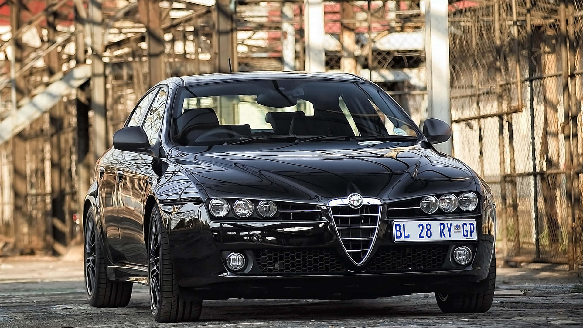 Alfa Romeo 159 桌布 壁紙 — 一輛時尚的黑色愛快羅密歐跑車在日光下閃耀，身後是工業小巷和生鏽的金屬結構。