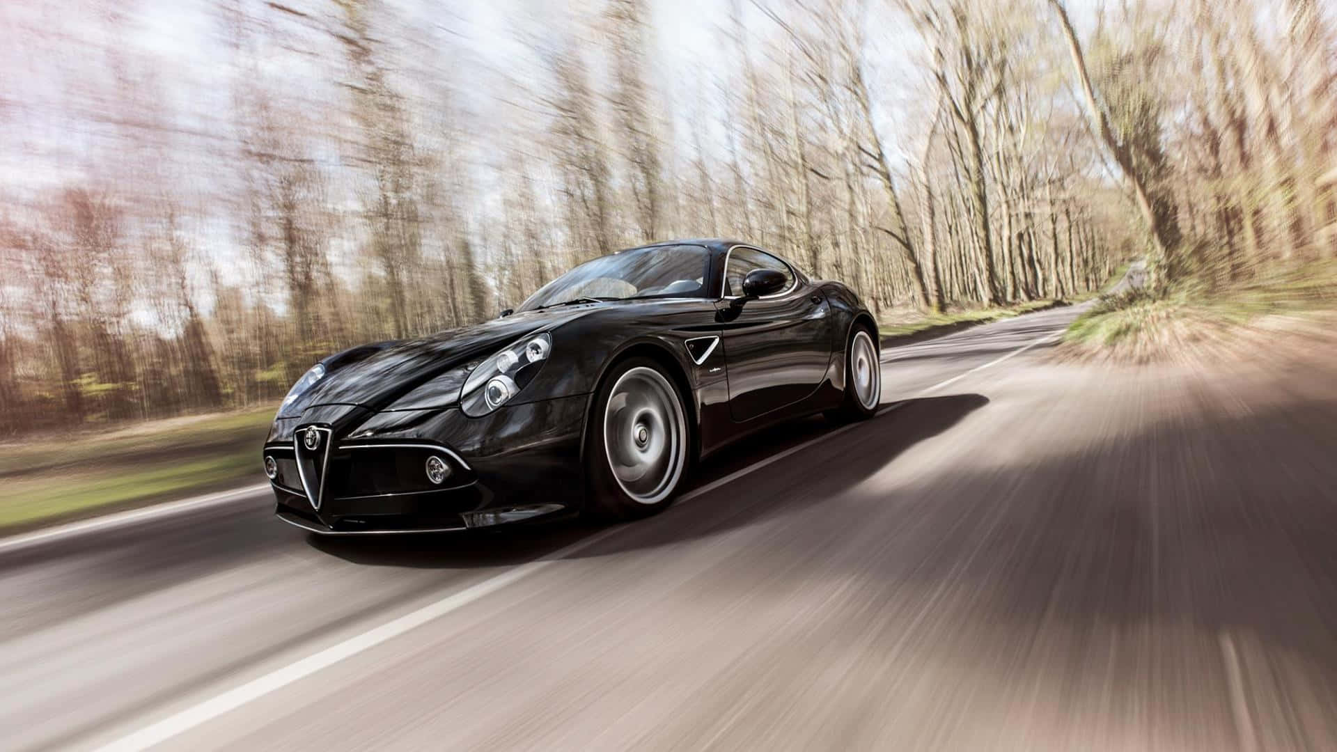 Alfa Romeo 8c Competizione 桌布 壁紙 — 一輛時尚的黑色愛快羅密歐跑車在蜿蜒的林間道路上飛馳，樹木模糊，陽光穿過光禿的樹枝灑落。