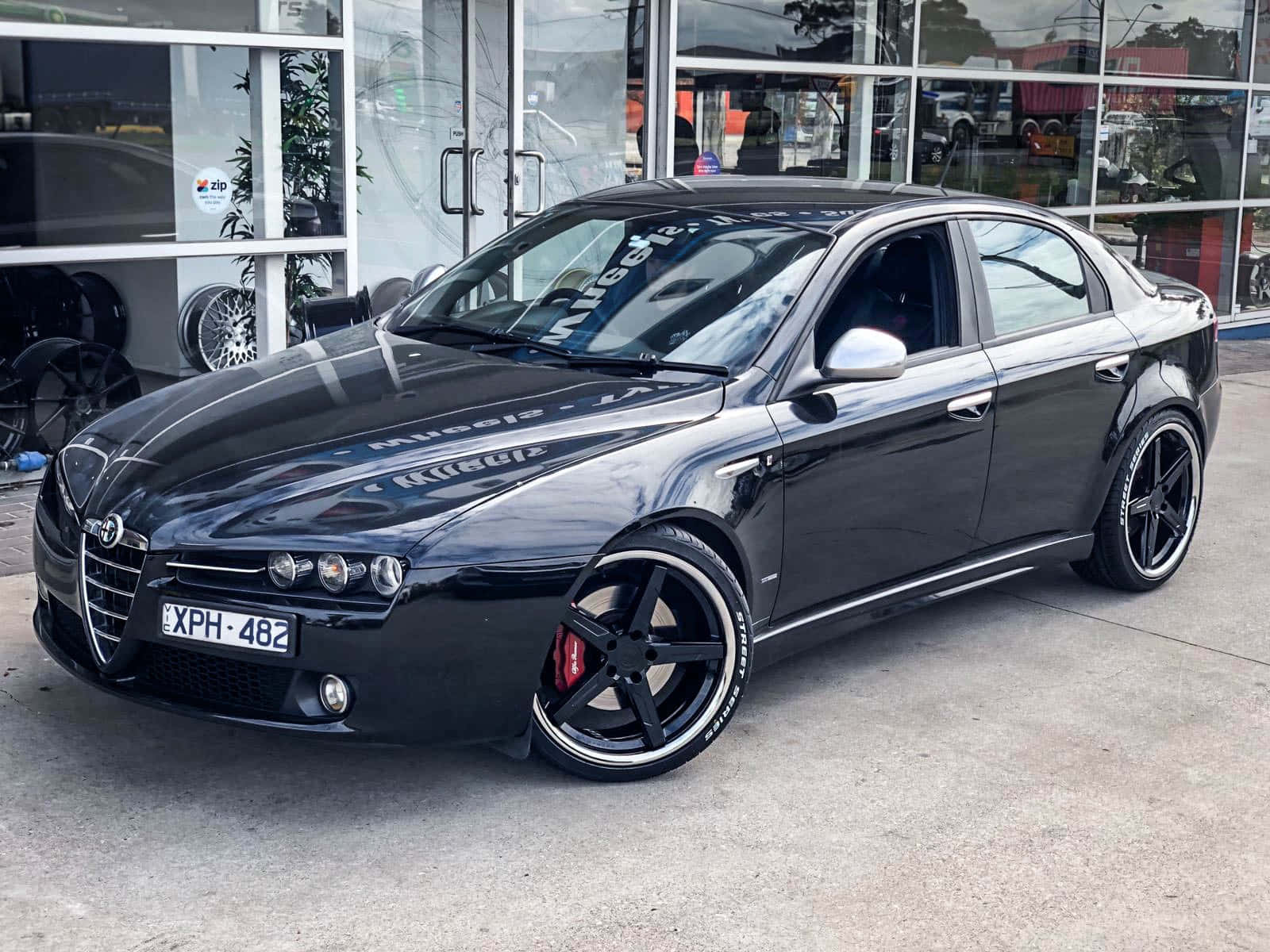 Alfa Romeo 159 桌布 壁紙 — 一輛時尚的黑色愛快羅密歐轎車，配備定制輪圈和紅色制動卡鉗，停在玻璃幕牆的經銷商門外。