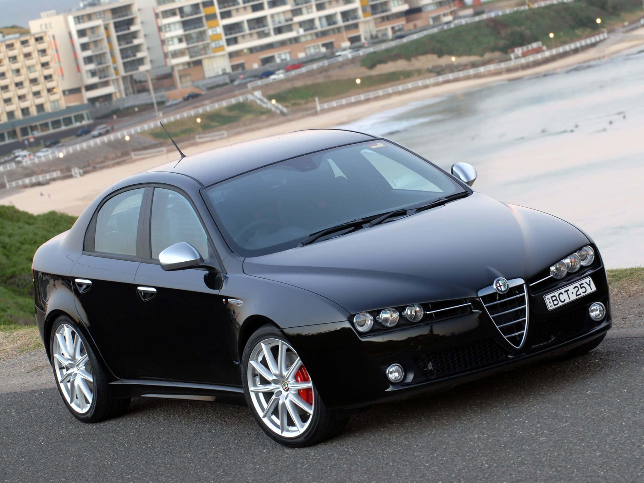Alfa Romeo 159 桌布 壁紙 — 一輛時尚的黑色愛快羅密歐轎車，配備銀色輪圈和紅色制動卡鉗，停泊在俯瞰沙灘和城市建築的海岸公路上。