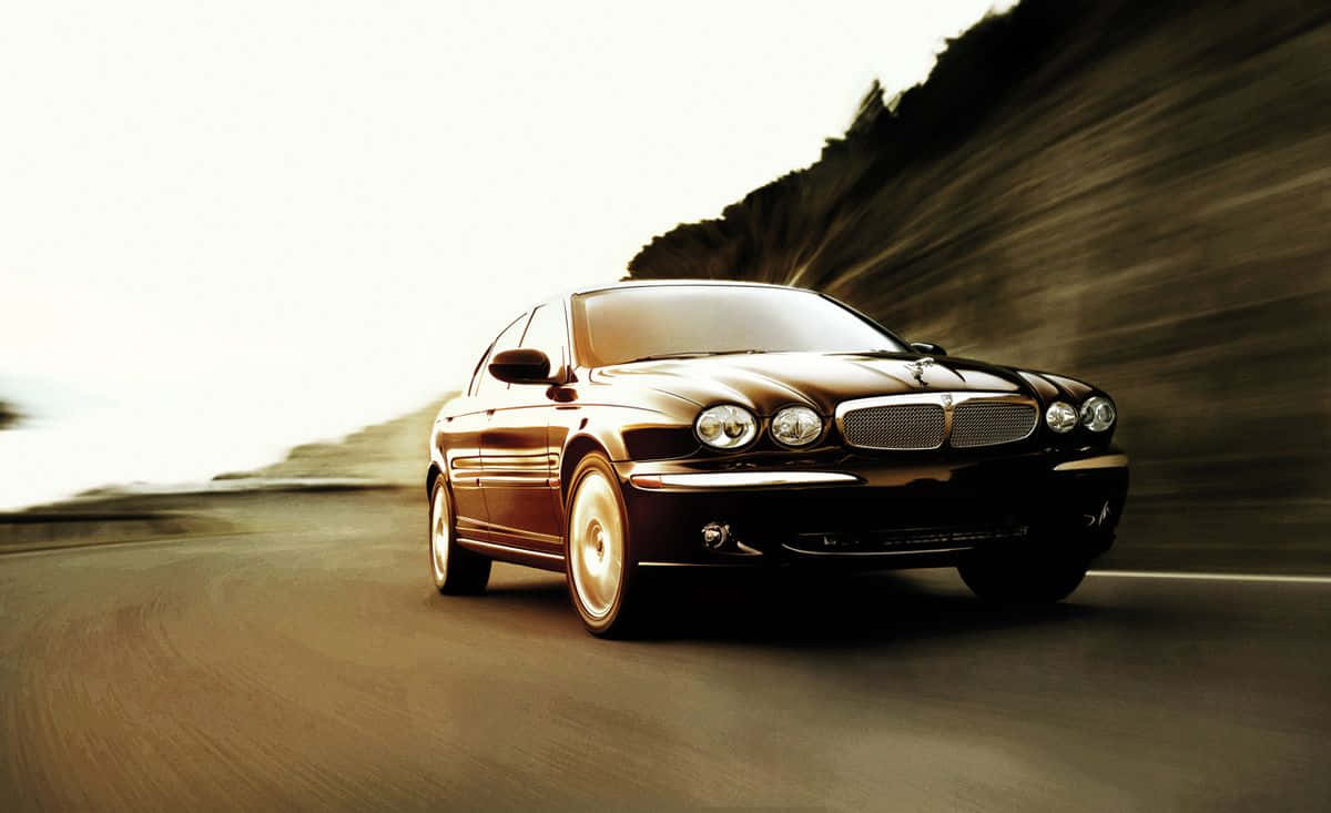 Jaguar X-type 桌布 壁紙 — 一輛時尚的黑色捷豹房車沿著蜿蜒的海岸道路飛馳，動態模糊效果強調了速度。