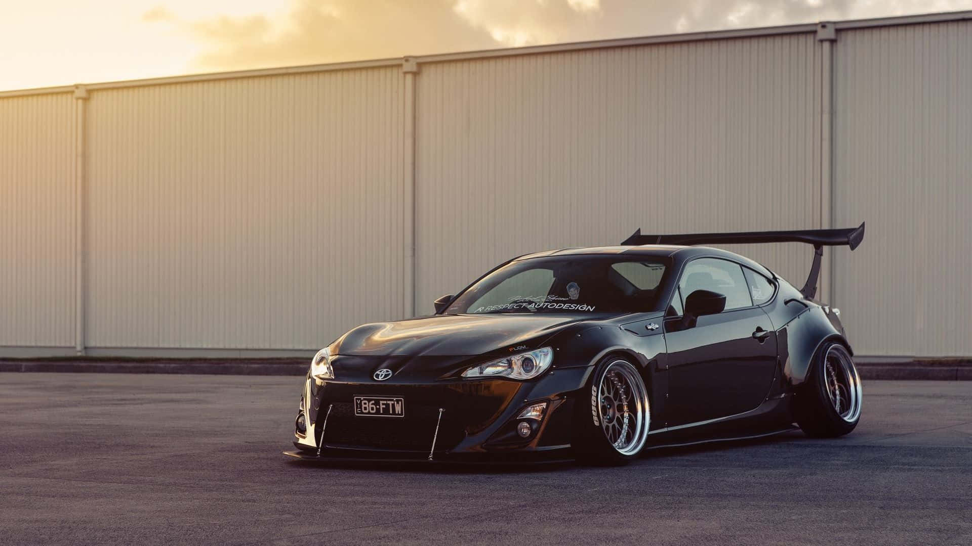 Toyota 桌布 壁紙 — 一輛時尚的黑色改裝跑車，帶有大型後翼，停在日落時分的米色工業牆前。