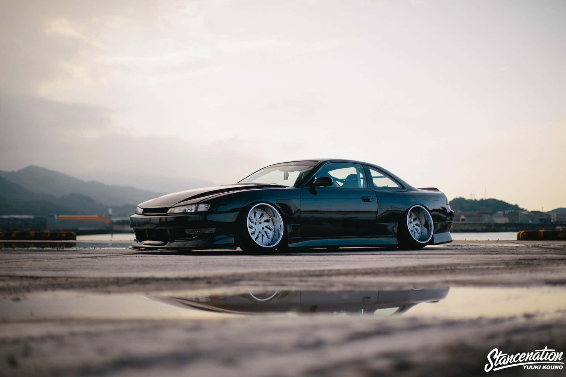 Stance Jdm 桌布 壁紙 — 一輛時尚的黑色改裝跑車配銀色輪圈，低趴於濕漉漉的路面上，在暮色中映襯著朦朧的山景倒影。