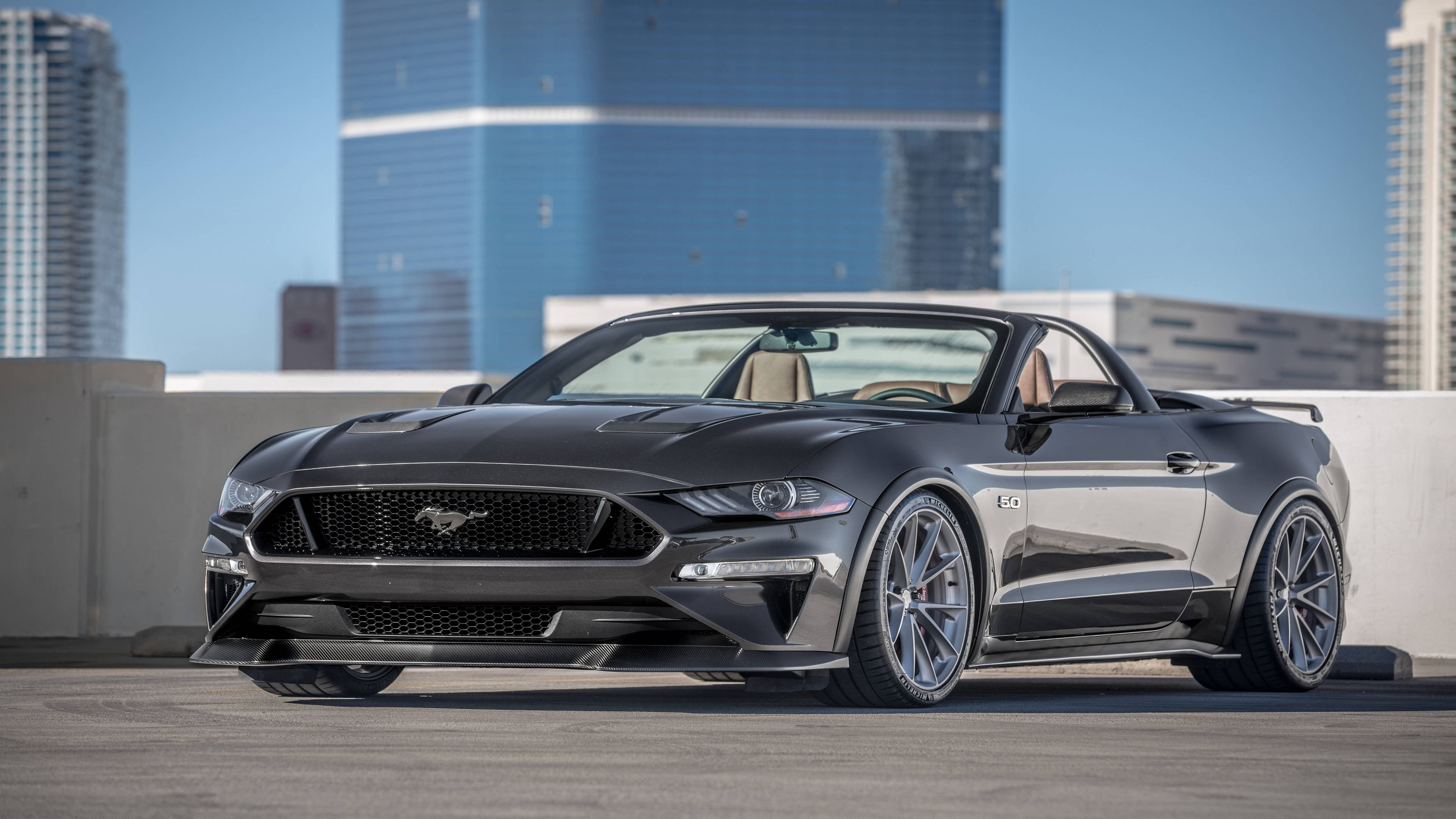 mustang HD 桌布 壁紙 — 一輛時尚的黑色敞篷跑車停放在屋頂上，背景是現代化的摩天大樓，天空清澈湛藍。