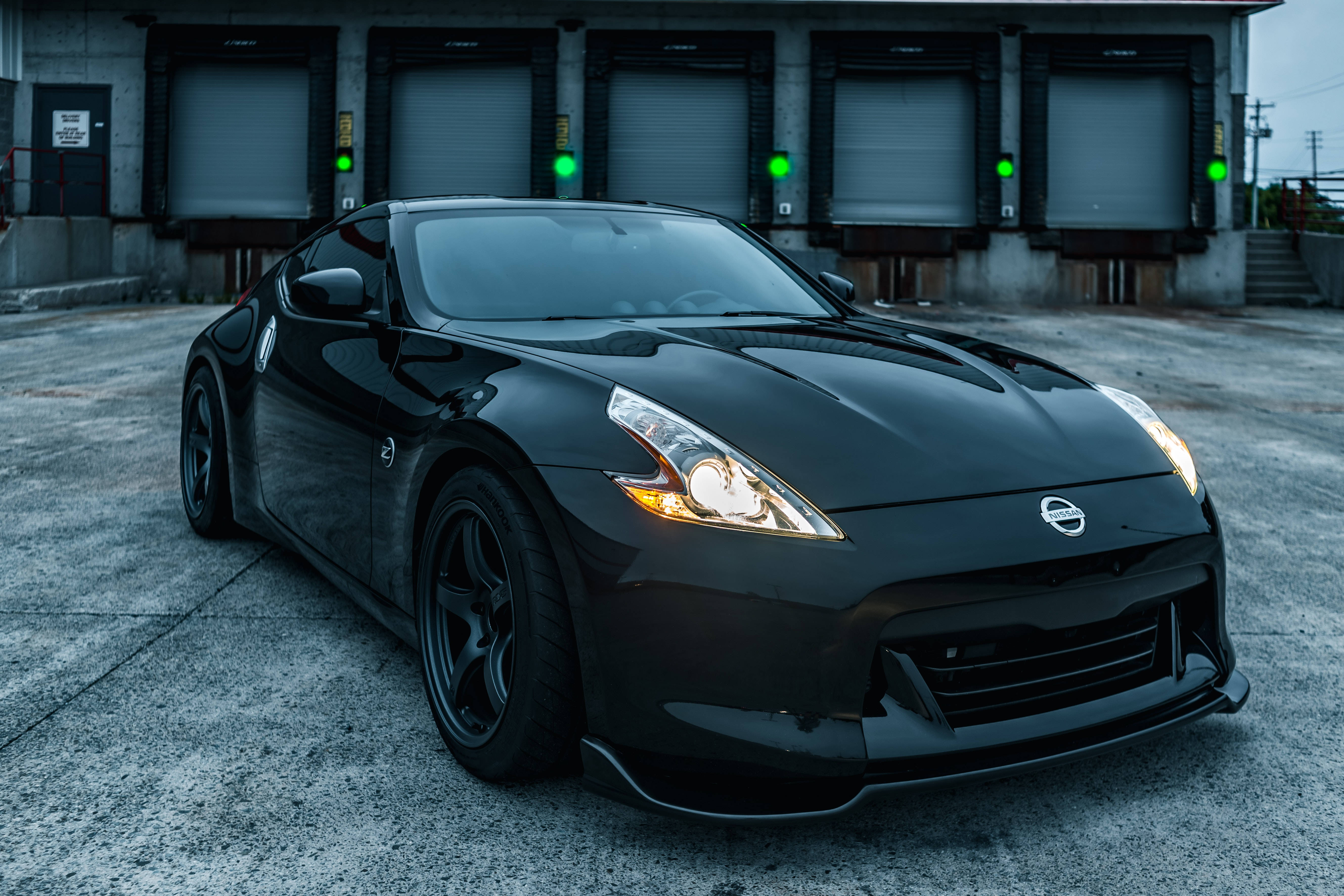 Nissan 桌布 壁紙 — 一輛時尚的黑色日產370Z跑車在暮色中閃耀，車頭燈亮著，停在工業車庫門前。
