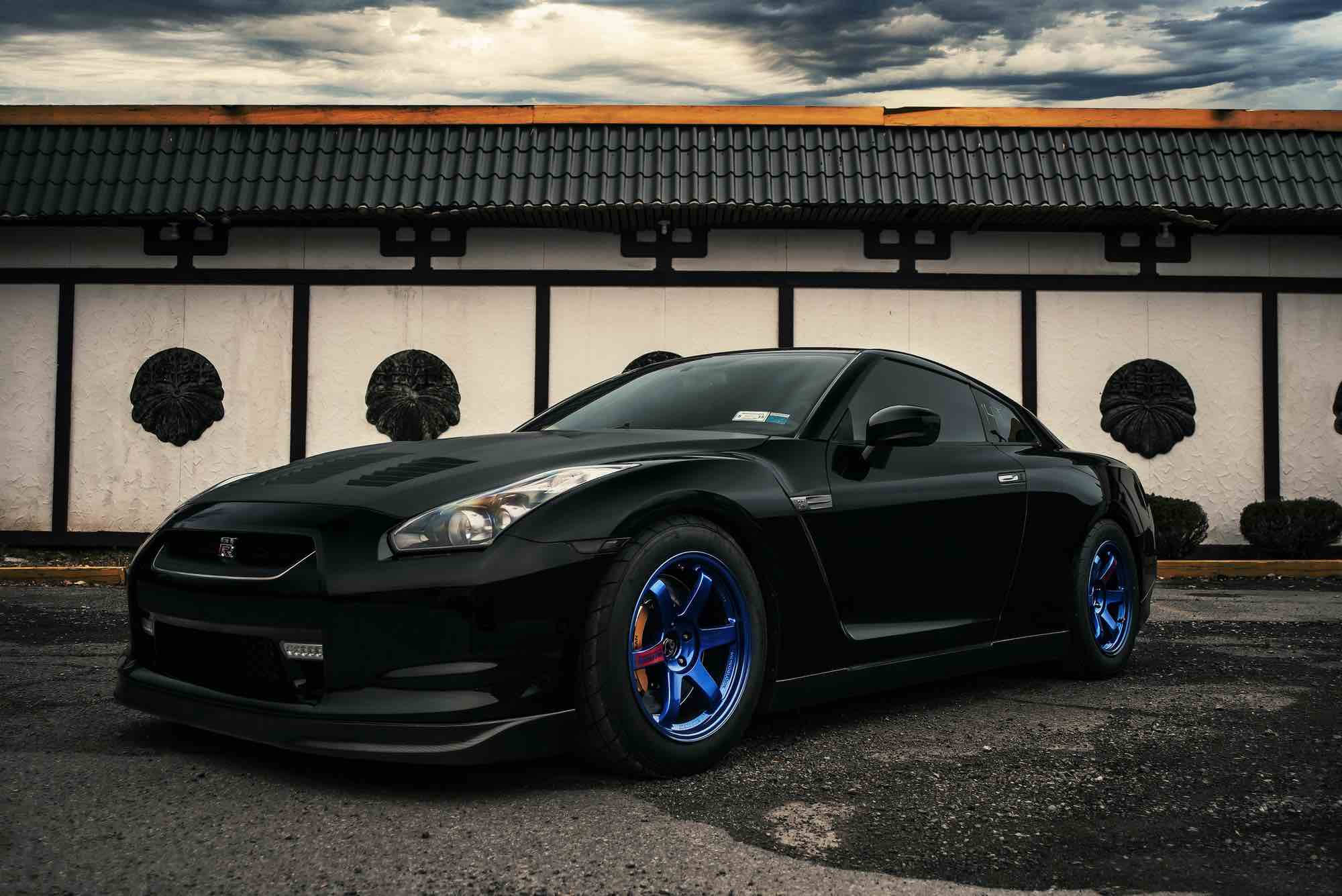 桌布 壁紙 — 一輛時尚的黑色日產GT-R，配以鮮藍色輪圈，停泊在傳統日式建築前，天空陰沉。