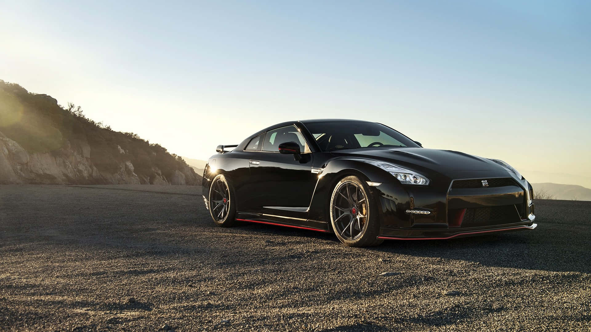 Gtr 桌布 壁紙 — 一輛時尚的黑色日產GT-R跑車，帶有紅色點綴，停泊在日落時分的碎石路上，背景是山丘。