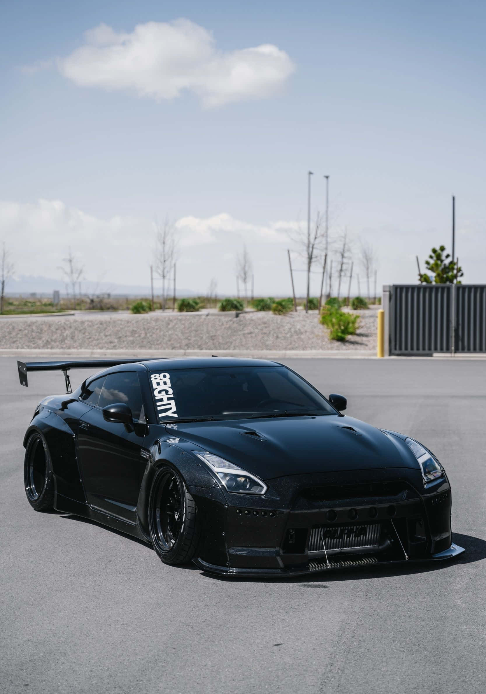 Nissan 桌布 壁紙 — 一輛時尚的黑色日產GT-R跑車，帶有大型後尾翼，停在瀝青停車場上，天空局部多雲。
