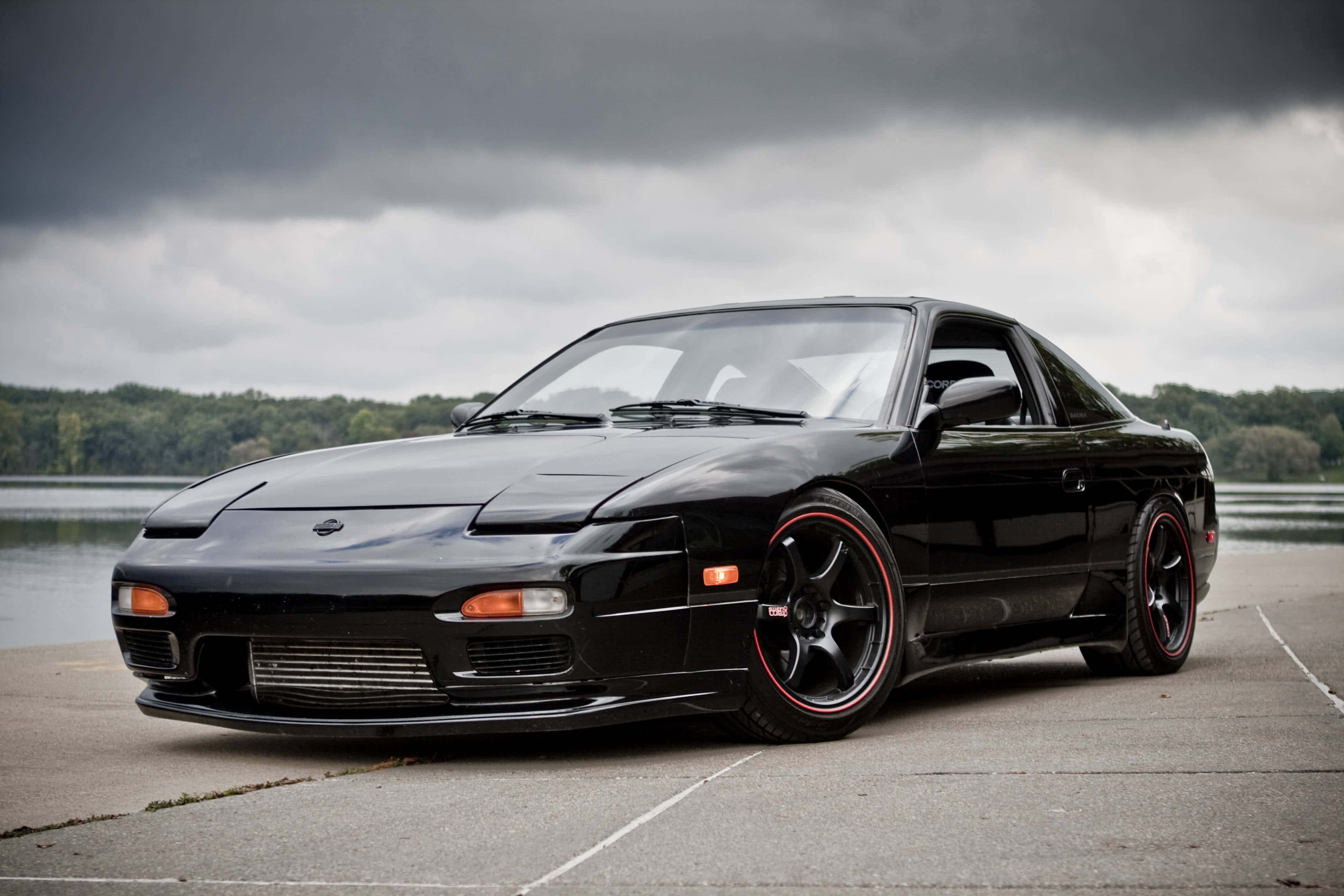 Nissan 180sx 桌布 壁紙 — 一輛時尚的黑色日產S14跑車，配備紅色輪圈，停放在濕漉漉的混凝土場地上，背景是暴風雨前的灰色天空、湖泊和森林。